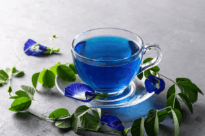A clear glass teacup filled with vibrant blue butterfly pea flower tea sits on a matching saucer, surrounded by fresh green leaves and vivid blue Clitoria ternatea blossoms. Set against a soft gray background, the composition highlights the tea’s natural color and cultural allure—Thailand’s magical drink known for its dramatic color shift when mixed with citrus.
