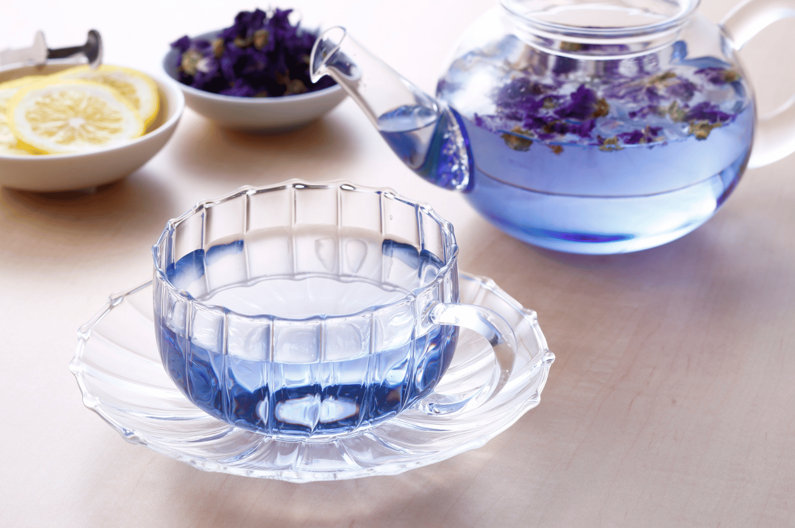 Butterfly-Tea- (2) Glass teacup and teapot filled with vivid blue butterfly pea flower tea, with floating purple petals hinting at its botanical infusion. Nearby, lemon slices and fresh blossoms rest in white dishes, suggesting the citrus transformation that turns the tea from deep blue to vibrant violet. The serene tabletop setting evokes Thailand’s ritualistic charm and the visual magic of this color-changing herbal drink.