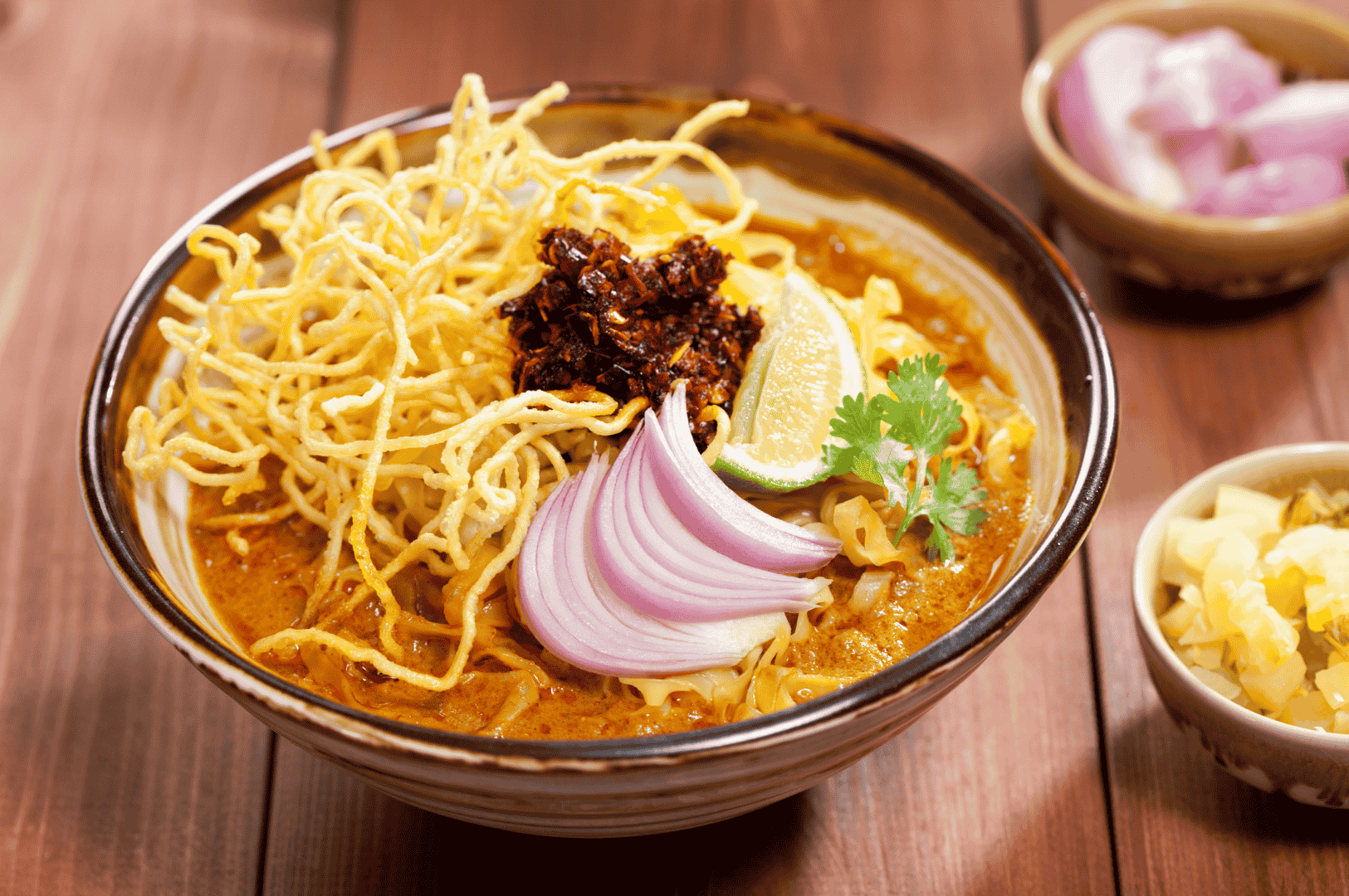 Khao-Soi-1 A ceramic bowl of Khao Soi, Northern Thailand’s iconic curry noodle soup, brimming with creamy coconut curry broth and layered with both boiled and crispy egg noodles. Garnished with a nest of golden fried noodles, a dollop of chili paste, fresh cilantro, red onion slices, and a lime wedge, the dish is surrounded by small side bowls of pickled mustard greens and extra onions. Set on a rustic wooden surface, the vibrant textures and colors evoke the warmth, complexity, and cultural depth of traditional Thai cuisine.