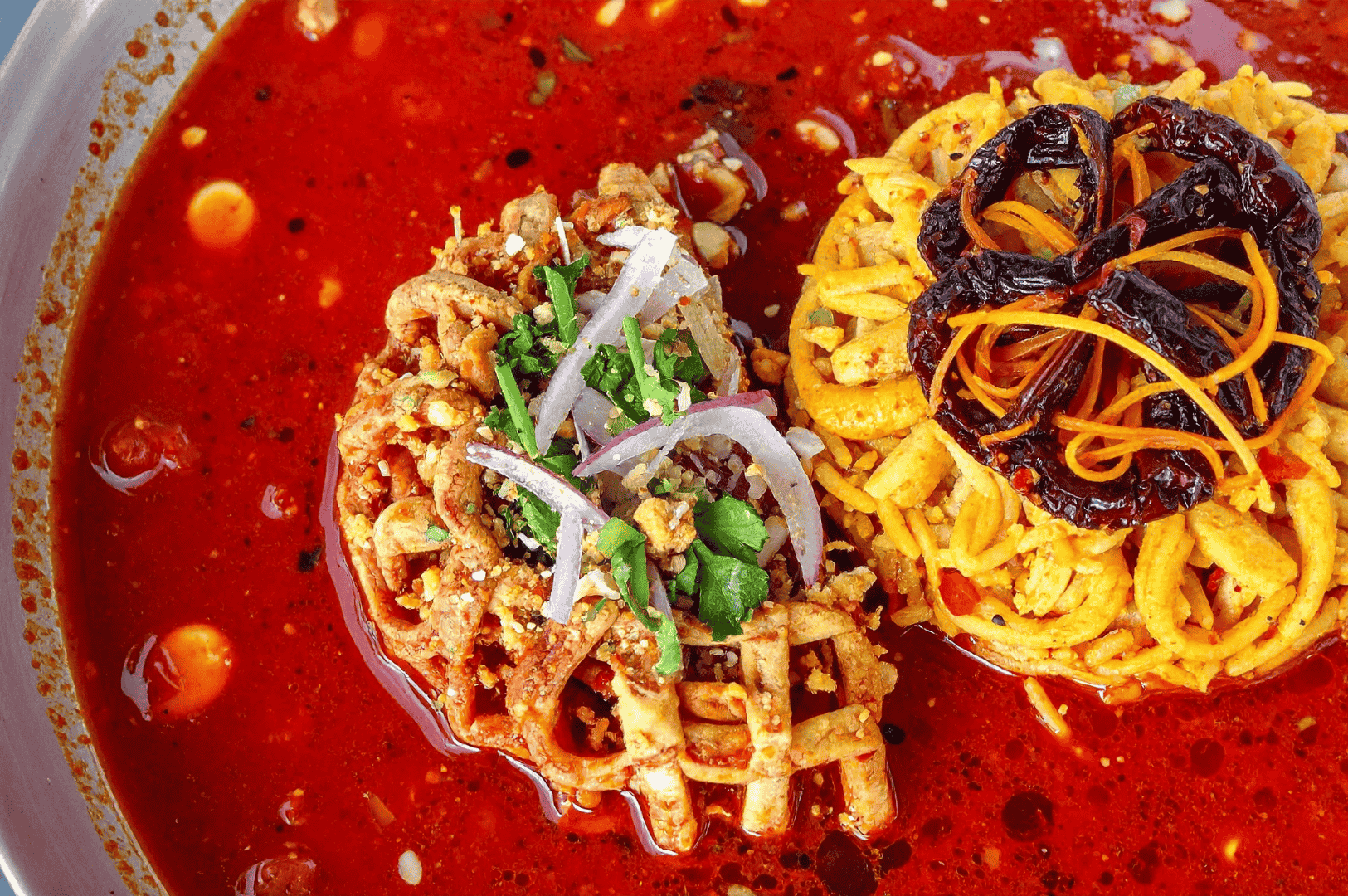 Khao-Soi-2 Close-up of Khao Soi, Northern Thailand’s iconic curry noodle soup, showcasing a vibrant red curry broth with crispy and soft egg noodles. The left portion is topped with minced meat, chopped red onions, cilantro, and green onions, while the right side features dried red chilies and thinly sliced carrots. Chili oil glistens on the surface, enhancing the dish’s bold flavor profile. Served in a rustic bowl, the contrasting textures and vivid colors celebrate the depth and diversity of Thai culinary tradition.