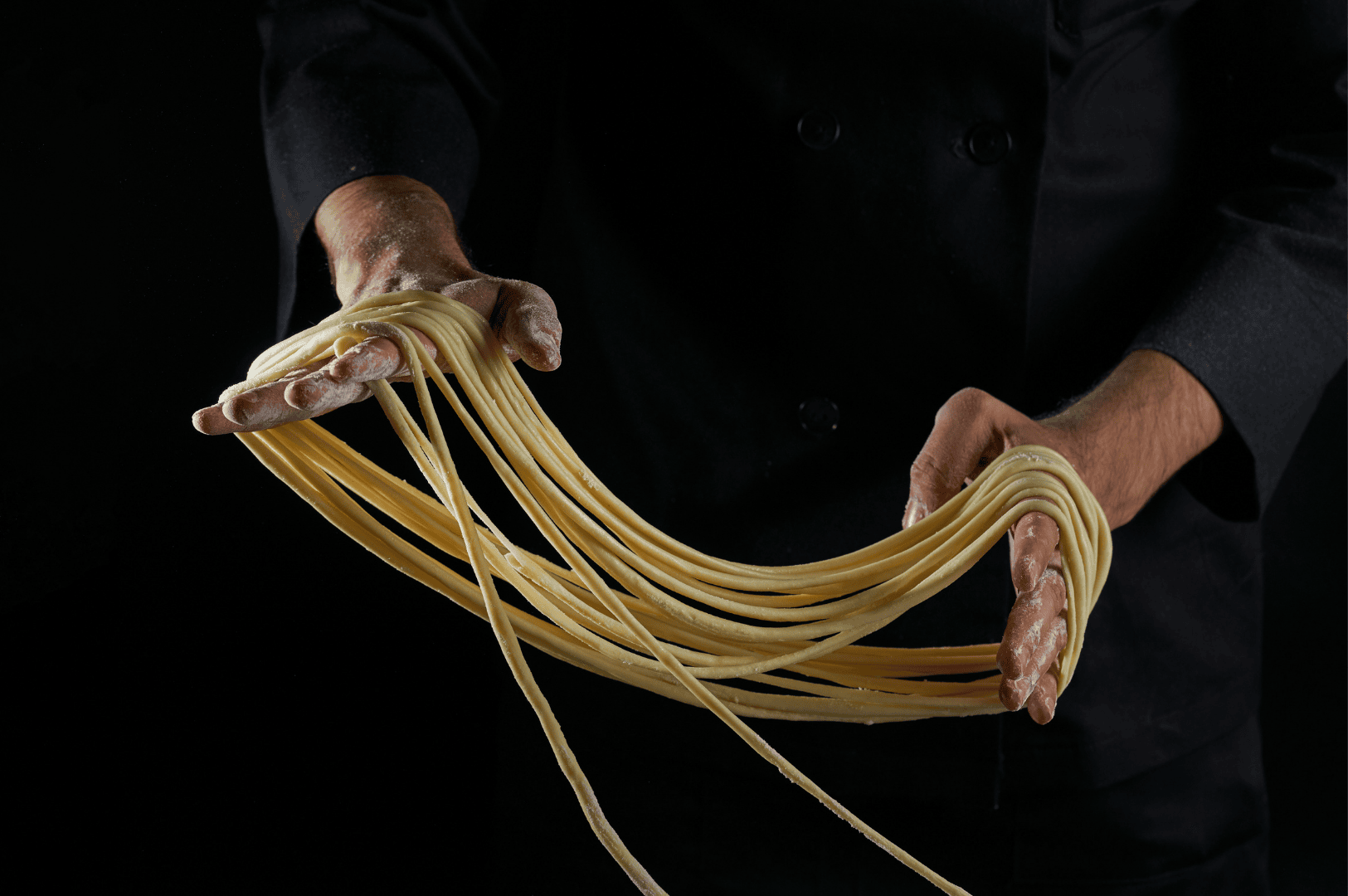 Khao-Soi-4 A dramatic close-up of fresh egg noodles being hand-stretched by a chef dressed in a dark uniform, set against a black background. The long, elastic strands evoke the artisanal craftsmanship behind Khao Soi, Northern Thailand’s iconic curry noodle soup. The image highlights the tactile beauty and traditional technique of noodle preparation, underscoring the dish’s cultural depth and culinary artistry.