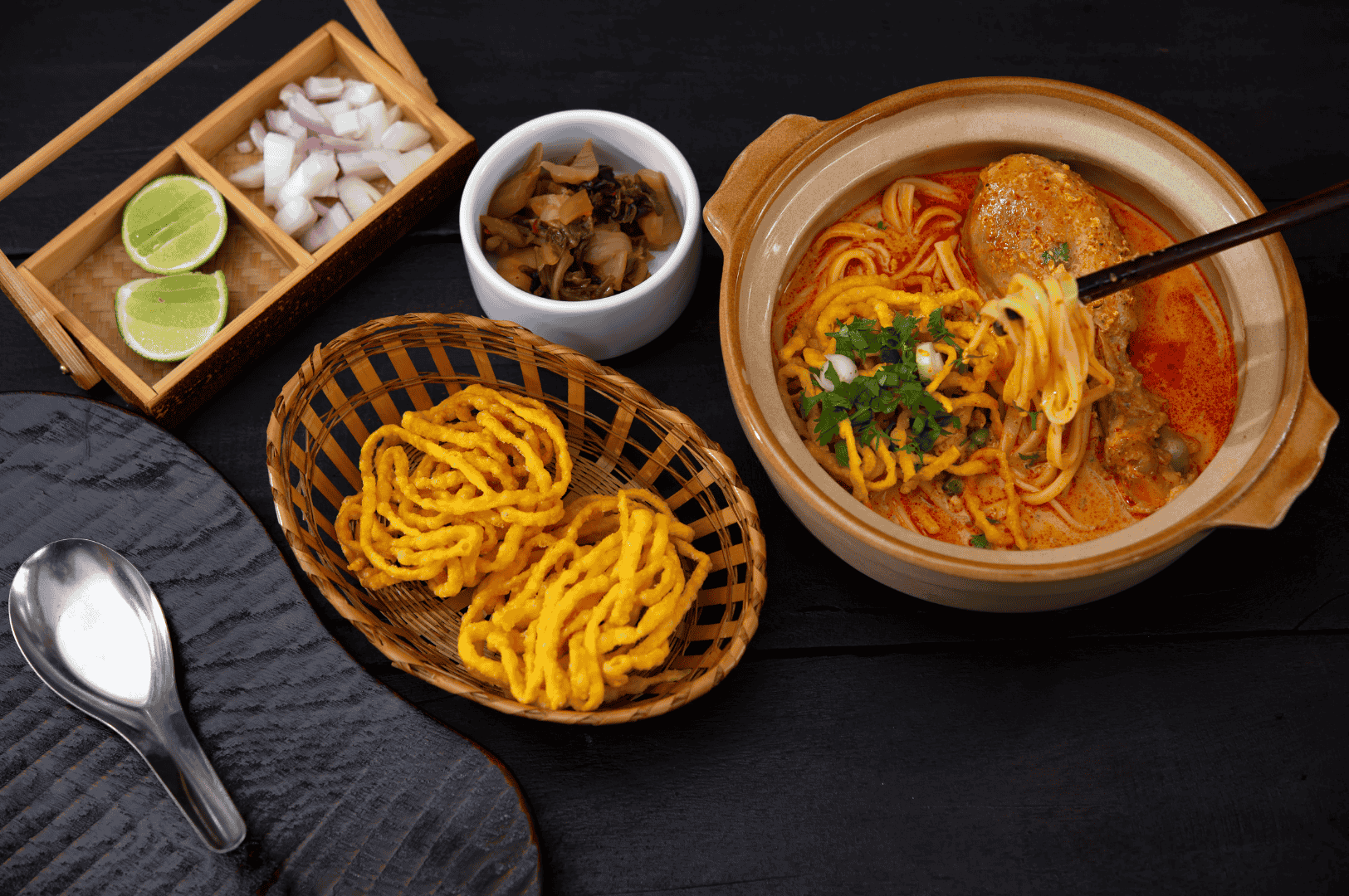 Khao-Soi-5.5 Bowl of Khao Soi, Northern Thailand’s signature coconut curry noodle soup, featuring yellow egg noodles, a braised chicken drumstick, crispy fried noodle topping, and fresh herbs like cilantro and green onion. Served with traditional sides—lime wedges, diced red onion, pickled mustard greens, and extra crispy noodles—on a rustic wooden tray, capturing the vibrant flavors and textures of this iconic regional dish.