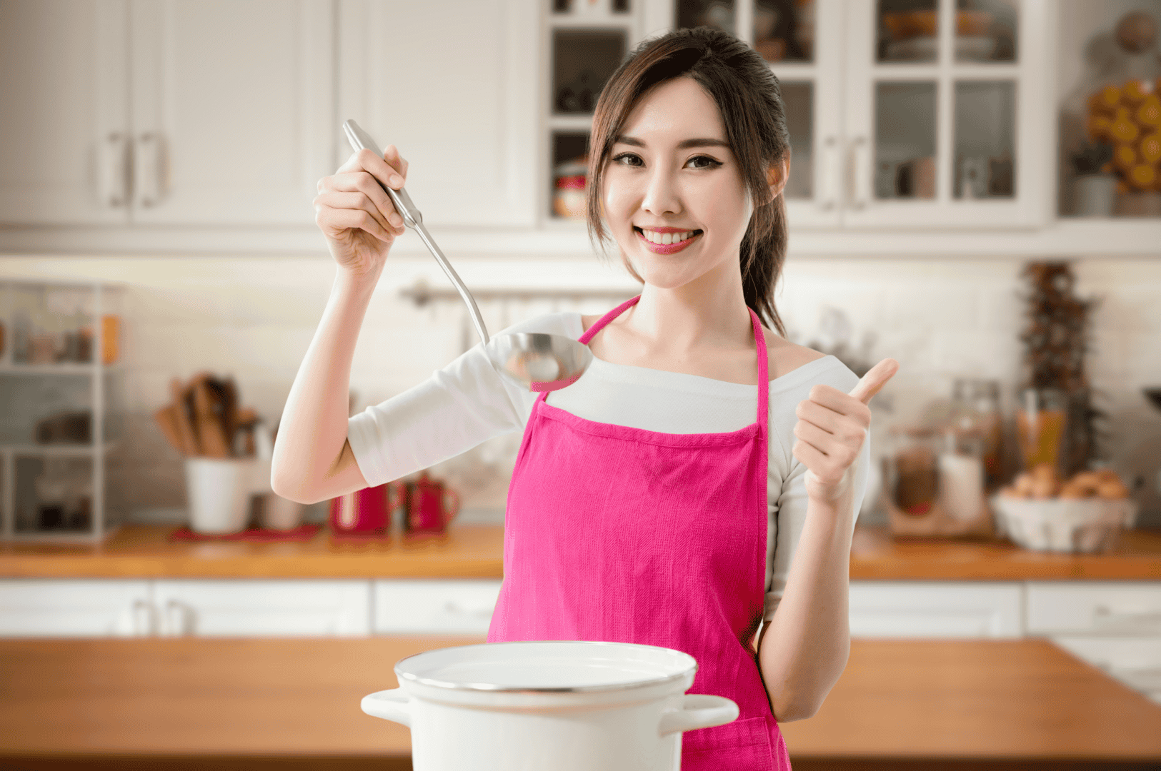 Khao-Soi-6 Smiling cook in a bright pink apron giving a thumbs-up while preparing food in a modern kitchen, holding a ladle above a white pot. The cheerful scene evokes the warmth and hospitality behind Northern Thailand’s beloved Khao Soi curry noodle soup, celebrating the joy of home cooking and regional culinary pride.