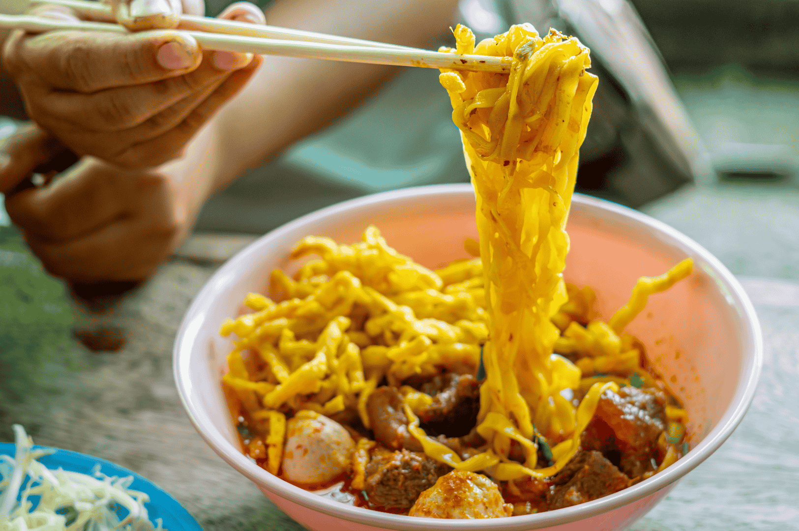 Khao-Soi-7 Hand lifting yellow egg noodles from a steaming bowl of Khao Soi, Northern Thailand’s iconic coconut curry noodle soup, featuring tender meat, fish balls, and a rich, reddish broth. Served with a side of shredded vegetables in a blue dish, the image captures the vibrant textures and bold flavors that define this beloved regional specialty.