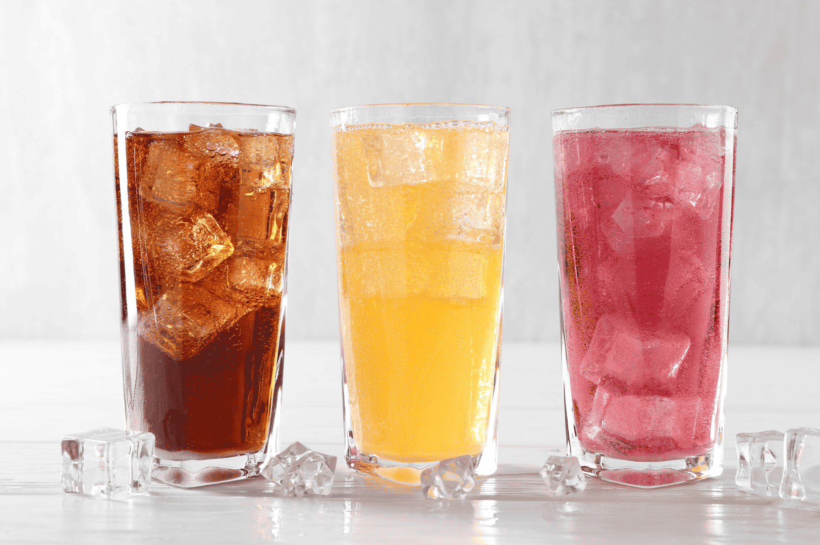 Khao-Soi-8 Three iced beverages in tall glasses—dark cola, bright orange soda, and pink fruit punch—set against a light background with scattered ice cubes. Though not traditional accompaniments, the colorful drinks offer a refreshing contrast to the rich, spicy flavors of Northern Thailand’s iconic Khao Soi curry noodle soup, reflecting the modern fusion of Thai dining experiences.