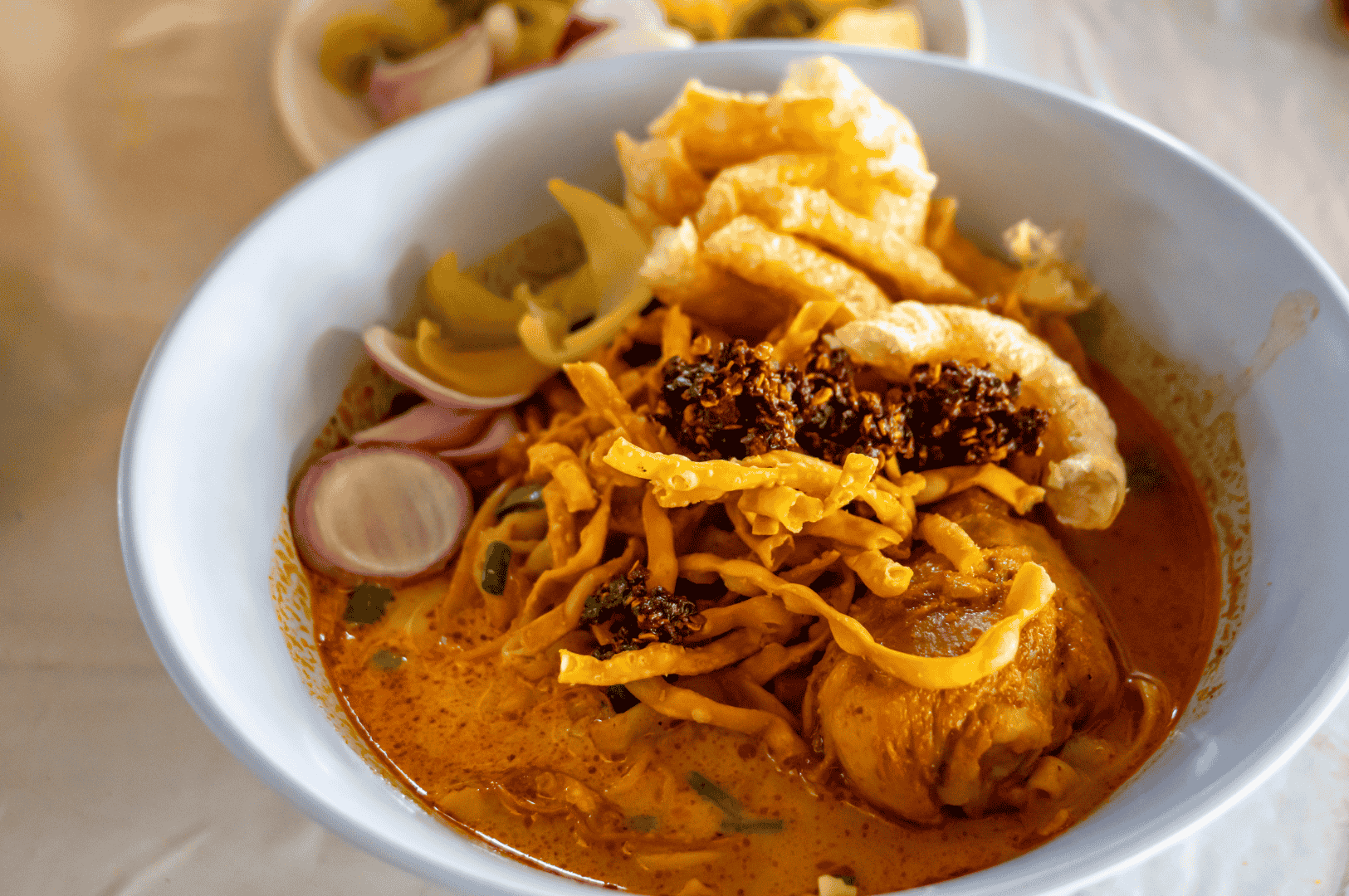 Khao-Soi-9 Bowl of Khao Soi, Northern Thailand’s iconic coconut curry noodle soup, featuring a chicken drumstick nestled in a creamy reddish-orange broth. Topped with crispy egg noodles, chili oil, pickled mustard greens, sliced shallots, and pork rinds, the dish showcases a vibrant mix of textures and bold flavors. A second bowl in the background hints at communal dining and the cultural richness of Thai cuisine.