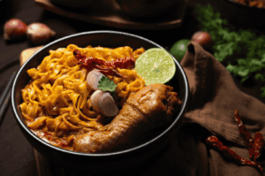 A steaming bowl of Khao Soi, Northern Thailand’s iconic curry noodle soup, featuring golden egg noodles submerged in rich coconut curry broth, topped with a tender chicken drumstick, crispy noodle garnish, sliced shallots, dried chilies, fresh cilantro, and a lime wedge. Presented in a black bowl on a rustic cloth napkin, surrounded by whole shallots and herbs, the dish evokes warmth, depth, and the vibrant flavors of Thai culinary tradition.