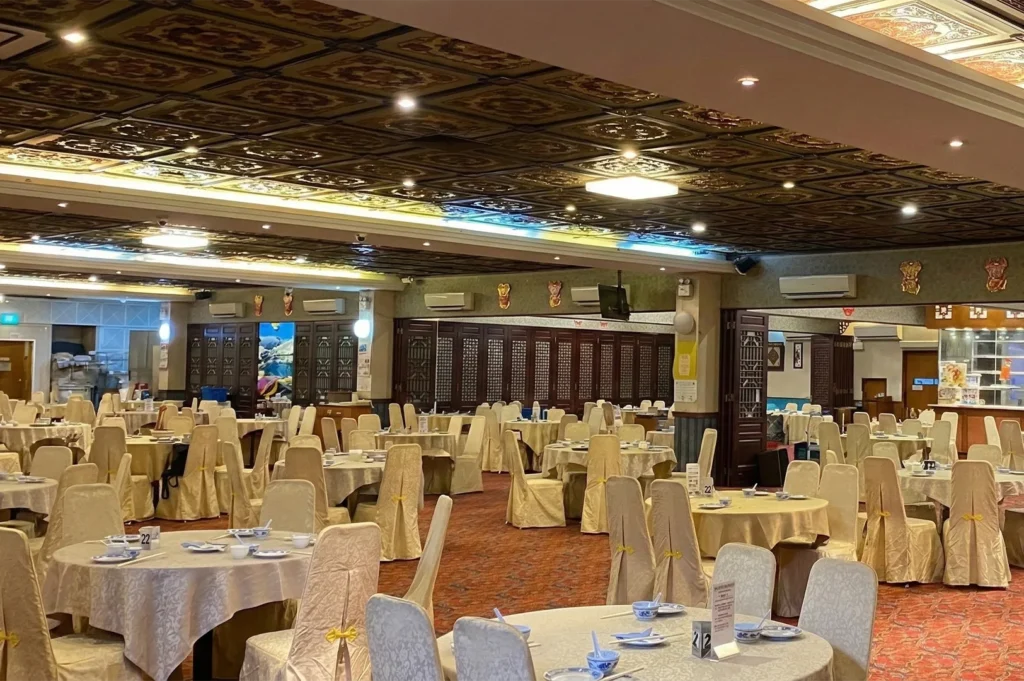 A large, traditional Chinese banquet hall with round tables covered in cream linens, gold-accented chairs, and a decorative coffered ceiling.