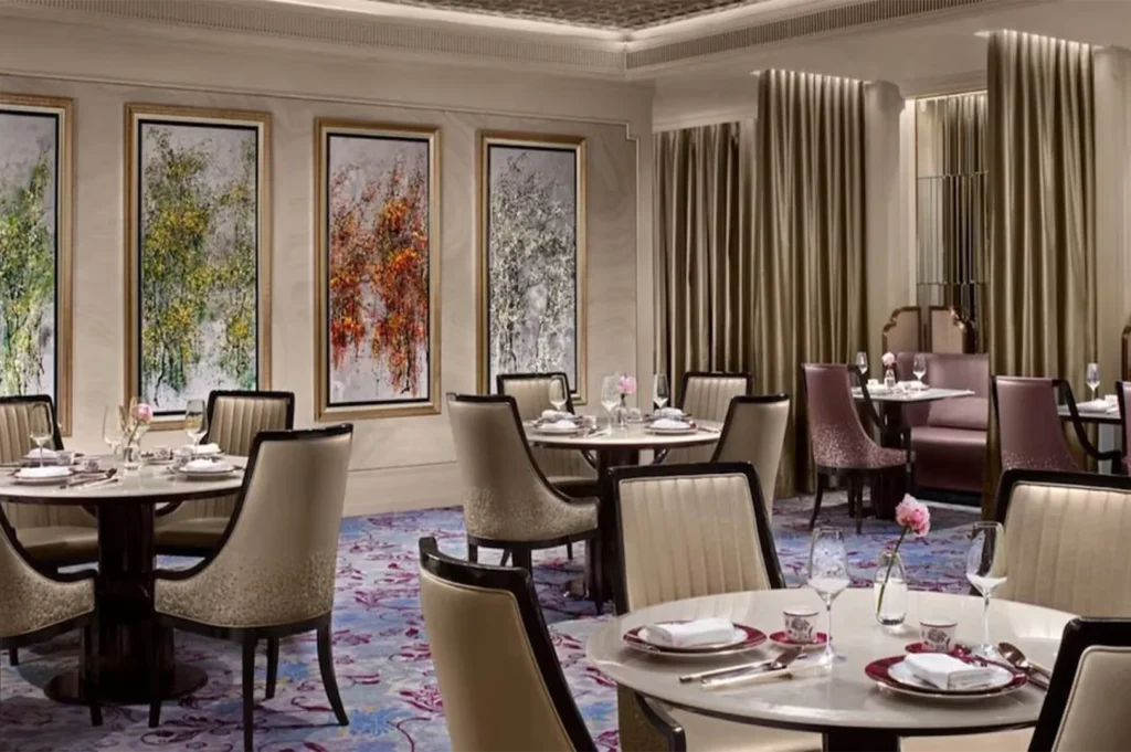 Upscale restaurant dining area featuring plush beige chairs, patterned blue carpets, and three large triptych floral paintings on the wall.