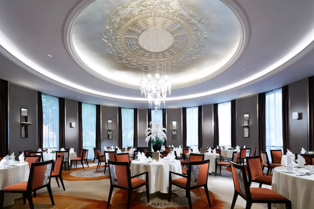 A high-end modern circular dining room with a grand chandelier, recessed ceiling lighting, and elegant orange-backed chairs.