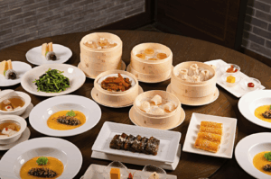 An abundant Cantonese dim sum spread arranged on a round wooden table, featuring bamboo steamers filled with assorted dumplings and shumai, alongside white plates of seaweed salad, sesame-coated rolls, and traditional desserts like jelly squares and layered sponge cakes—showcasing the rich diversity of dim sum beyond har gow and siu mai in an elegant, communal dining setting.