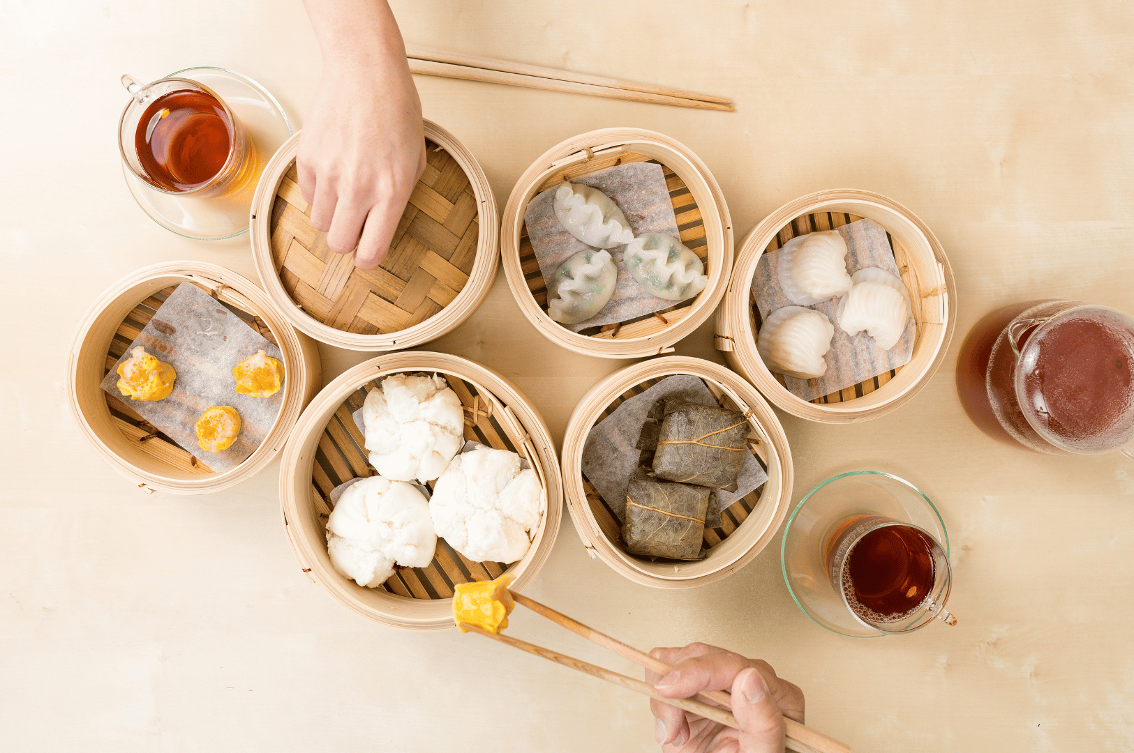 Cantonese-Dimsum- (2) Top-down view of a lively Cantonese dim sum table featuring seven bamboo steamers filled with a variety of dumplings, buns, and leaf-wrapped parcels—siu mai, translucent har gow, white bao, and more—accompanied by three glasses of tea and two hands actively serving, capturing the communal rhythm and rich diversity of dim sum beyond the classics