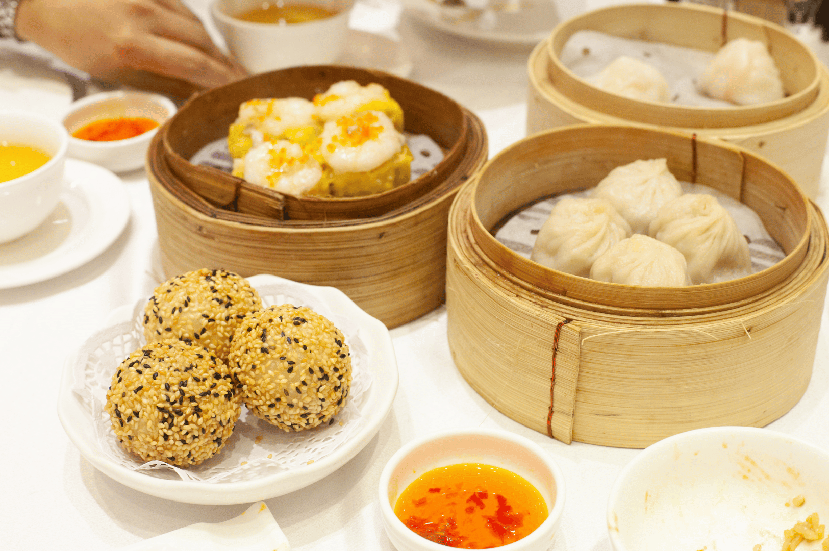 Cantonese-Dimsum- (3) Cantonese dim sum table set with bamboo steamers filled with siu mai topped with orange roe, pleated xiao long bao, and translucent har gow, alongside a plate of golden sesame balls and dipping sauces—capturing the vibrant textures, flavors, and shared rituals of dim sum dining beyond the familiar staples.