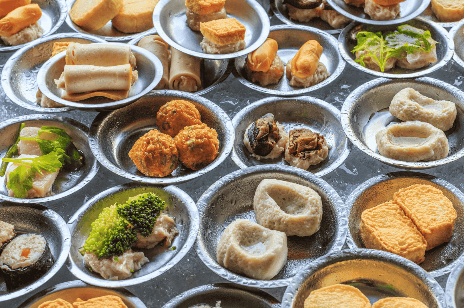 Cantonese-Dimsum- (4) Assorted Cantonese dim sum served in small metal plates—featuring steamed and fried delicacies like dumplings, spring rolls, tofu, and fish balls, garnished with broccoli and parsley—presented in a traditional style that celebrates the bite-sized diversity and sensory richness of dim sum beyond the usual favorites.