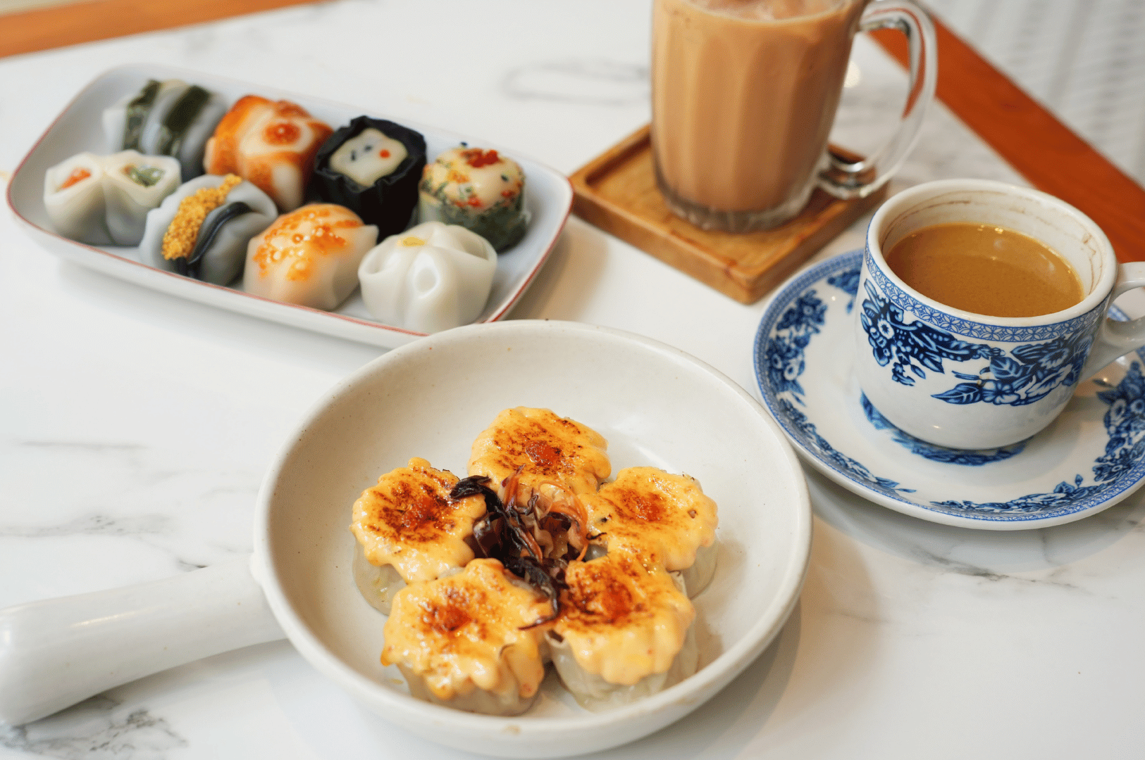 Cantonese-Dimsum- (6) Contemporary dim sum spread on a white marble table featuring six grilled or baked dumplings with charred creamy topping arranged in a circle, paired with a rectangular plate of colorful mochi-style rice cakes and two drinks—a glass mug of milk tea and a porcelain cup of coffee or tea—highlighting the fusion of savory and sweet in a visually refined Asian dining experience.