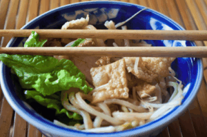 Overhead view of a bowl of Cao Lầu noodles, a Hoi An specialty with Japanese roots, featuring thick rice noodles, crispy fried tofu, fresh lettuce, and bean sprouts, served in a blue-and-white ceramic bowl with wooden chopsticks resting on top, set against a bamboo mat backdrop evoking traditional Vietnamese dining aesthetics.