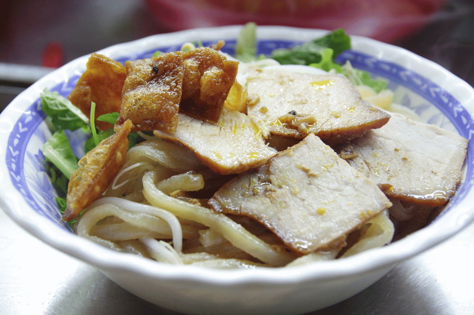 Cao-Lau- (4) Overhead shot of Cao Lầu noodles, Hoi An’s iconic dish with Japanese heritage, featuring thick rice noodles, roasted pork slices garnished with sesame seeds, crispy pork skin, and fresh herbs, served in a blue-and-white ceramic bowl—capturing the dish’s chewy texture, restrained seasoning, and historical fusion of Vietnamese and Japanese culinary traditions.
