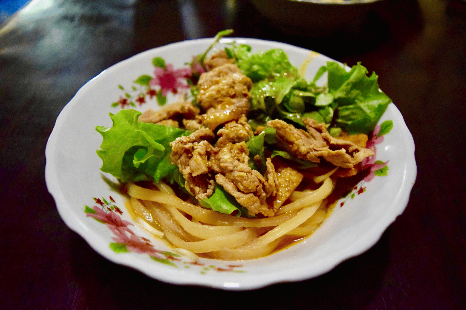 Cao-Lau- (5) Top-down view of a noodle dish featuring flat rice noodles, stir-fried meat slices, and crisp lettuce, served in a white bowl with floral patterns—highlighting the simplicity and freshness of everyday Vietnamese fare in contrast to the historical depth and chewy complexity of Hoi An’s Cao Lầu.
