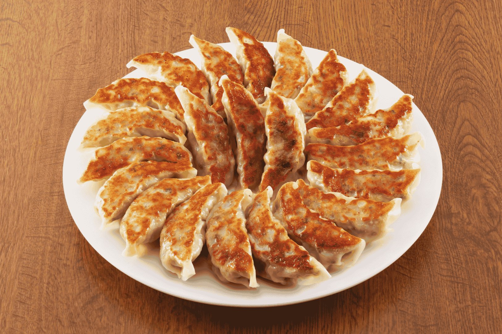 Century-Dumplings- (3) Golden-brown pan-fried dumplings arranged in a symmetrical circle on a white plate atop a wooden surface. Each dumpling bears crisp, caramelized edges and uniform pleats, showcasing the precision of time-honored techniques adapted for modern presentation. The circular layout evokes ritual and reverence, echoing the ancestral artistry behind every fold.