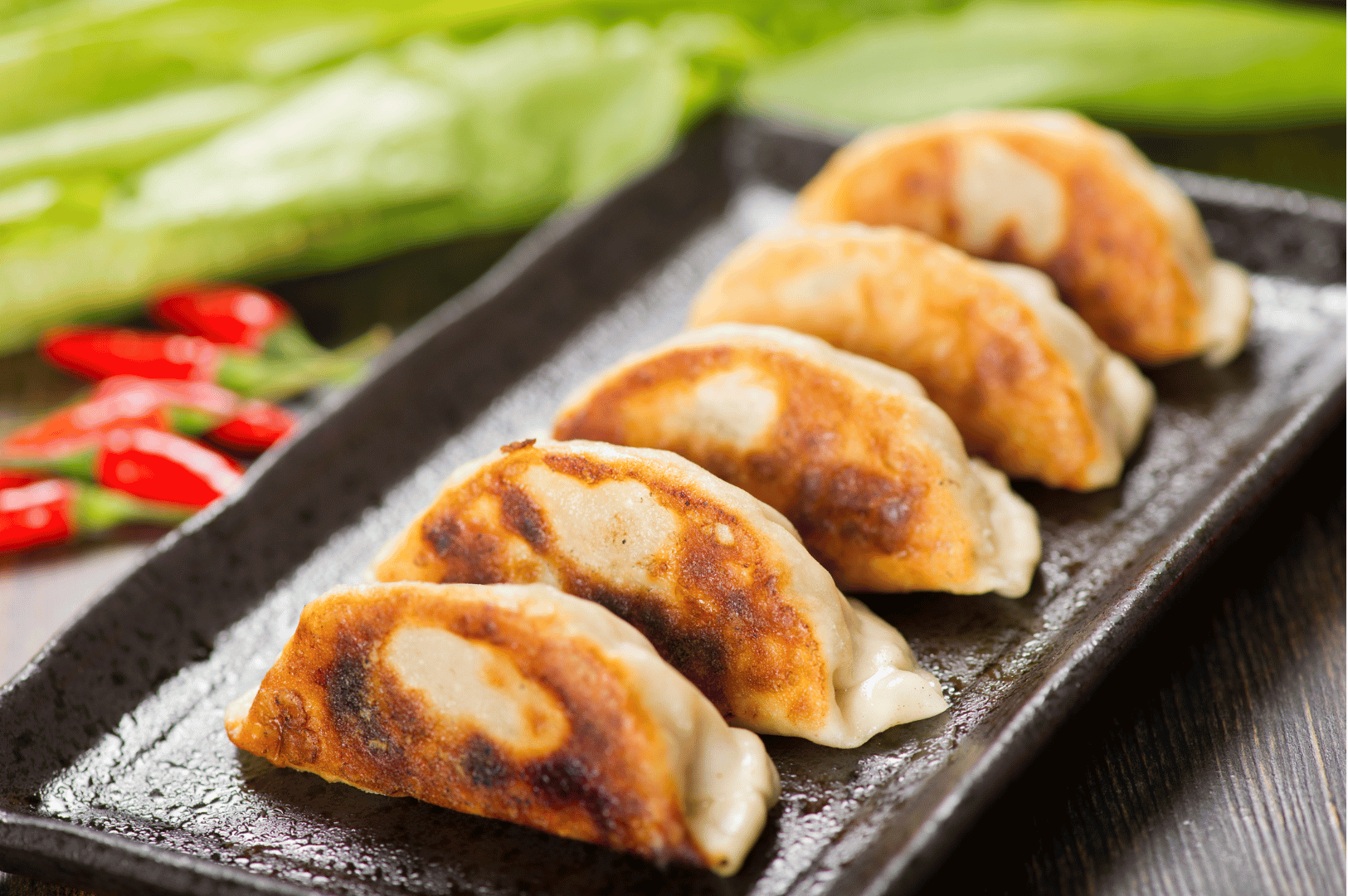 Century-Dumplings- (4) Five golden-brown pan-fried dumplings lined up on a sleek black plate, their crisp edges glistening with oil. In the softly blurred background, vibrant green vegetables and red chilies hint at the dish’s bold flavor profile. The composition evokes a modern mise en scène, where ancient dumpling-making rituals meet contemporary culinary styling.