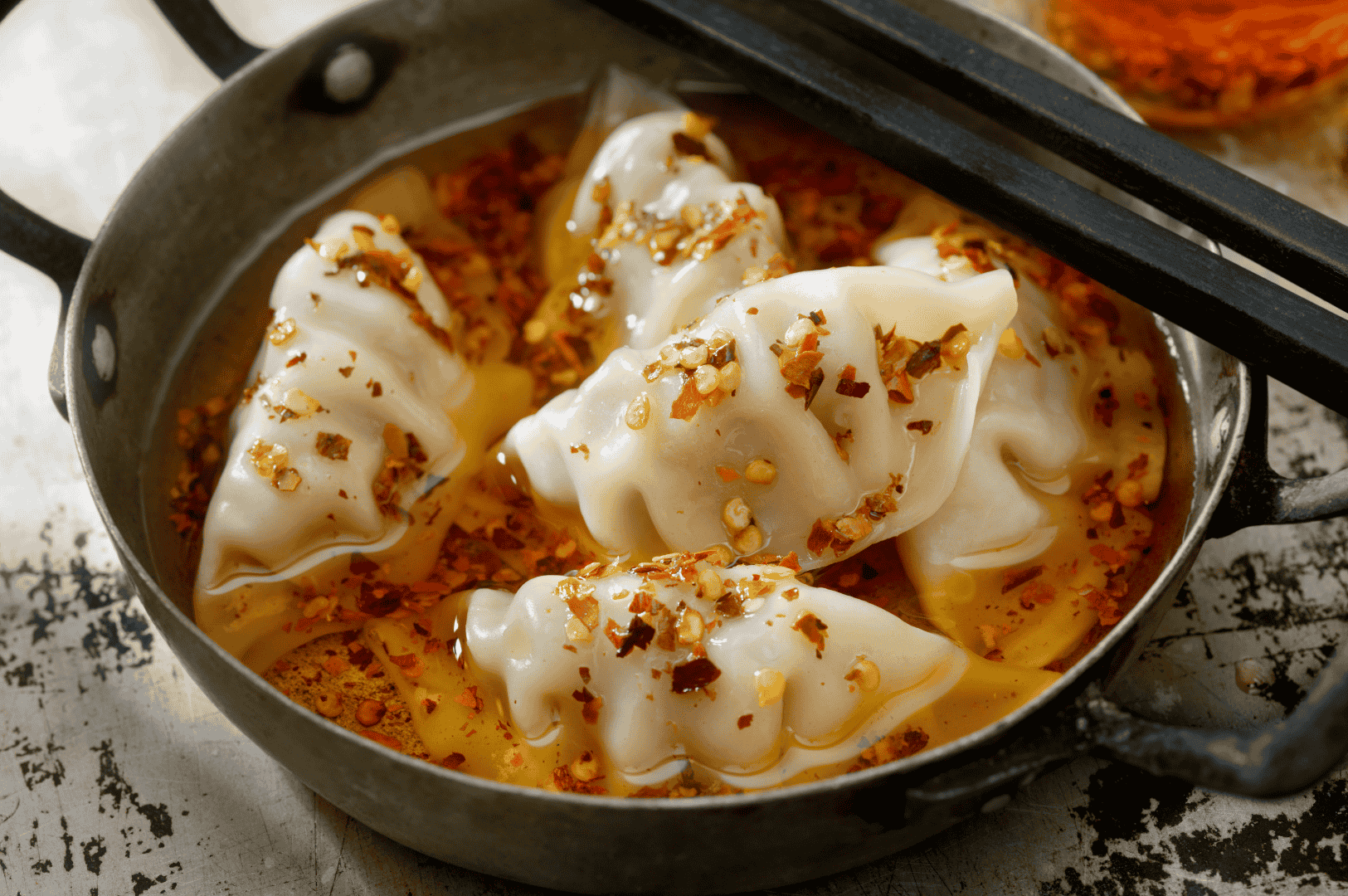 Century-Dumplings- (5) Steamed dumplings submerged in a vibrant red-orange chili broth, their translucent skins revealing savory fillings beneath. Crushed pepper flakes and chili oil swirl across the surface, hinting at bold, layered heat. A pair of black chopsticks rests on the pot’s edge, inviting the first bite. The rustic backdrop evokes a sense of home and heritage, where ancient flavors simmer with modern intensity.