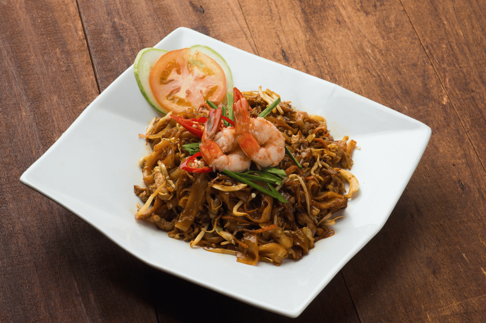 Elegant serving of Penang Char Kway Teow on a white square plate, showcasing flat rice noodles stir-fried with shrimp, bean sprouts, scallions, and red chili—garnished with fresh cucumber and tomato slices. The vibrant plating and rustic wood backdrop evoke the refined yet fiery soul of Malaysia’s iconic hawker dish.