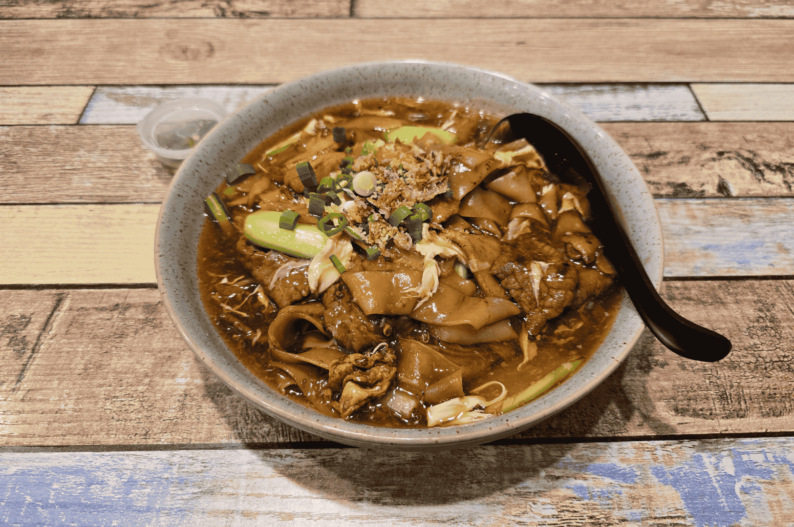 Bowl of Char Kway Teow, featuring silky flat rice noodles drenched in rich egg-laced gravy with sliced meat, scallions, and crispy shallots—served with a black soup spoon and chili condiment on a rustic multicolored wood table. A comforting counterpoint to Char Kway Teow’s fiery wok hei, this dish highlights Penang’s layered culinary heritage.