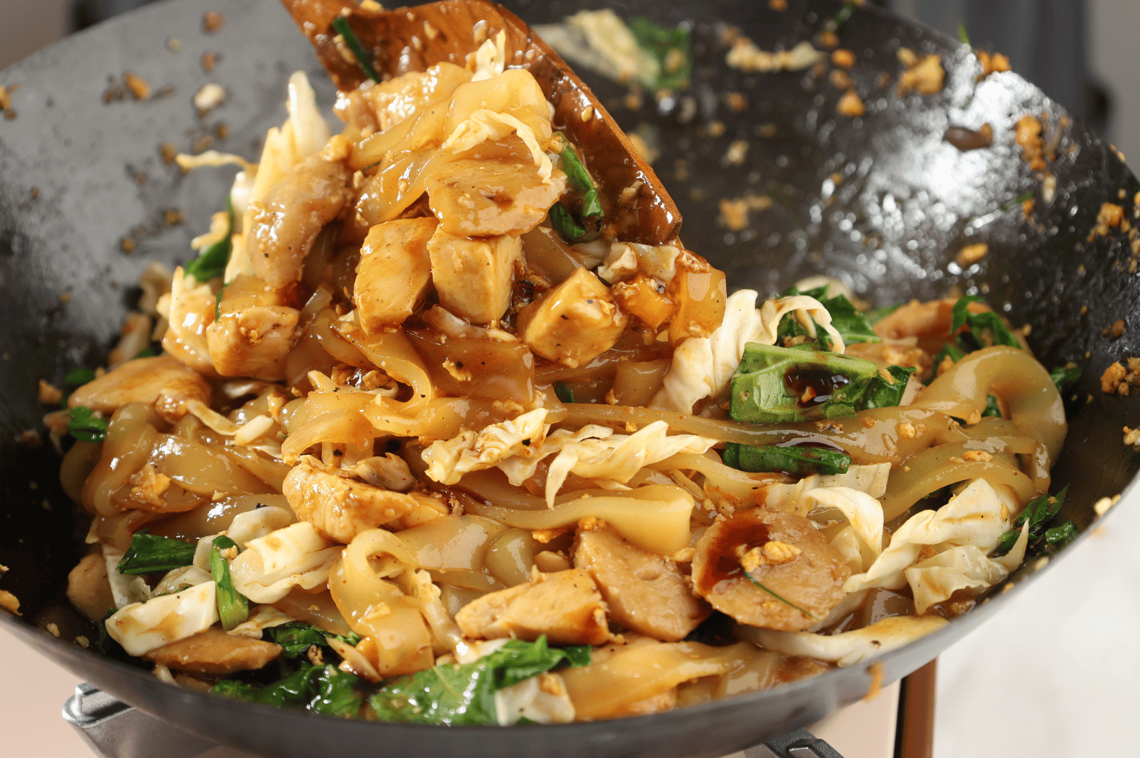 Close-up of stir-fried flat rice noodles sizzling in a seasoned wok, tossed with chicken, Chinese greens, and shredded cabbage in a dark soy glaze. Garlic remnants and sauce-stained edges evoke the high-heat intensity and tactile rhythm of street-side Char Kway Teow preparation.