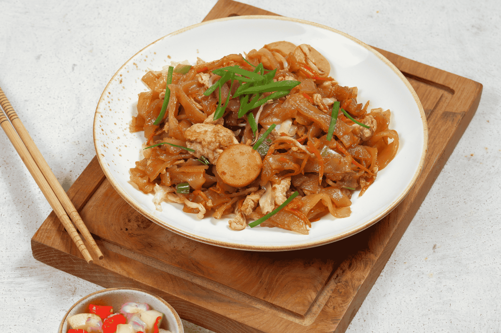 Artfully plated Char Kway Teow on a white dish with gold rim, featuring flat rice noodles stir-fried with fish cake slices, scrambled egg, bean sprouts, and scallions in a savory soy glaze—accompanied by pickled chili-onion relish and wooden chopsticks on a rustic board. A nod to Penang’s culinary elegance, where street food meets timeless tradition.