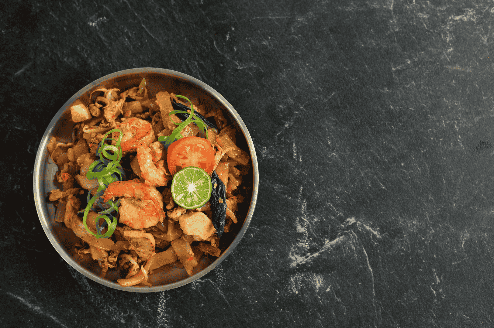 Colorful bowl of Char Kway Teow with shrimp, tofu, mushrooms, scallions, tomato slices, and lime—served on a dark textured surface. The bright garnishes and savory glaze evoke Penang’s coastal vibrancy, blending wok-fired depth with fresh, citrusy lift.