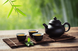 Black ceramic teapot and three matching teacups filled with amber-hued tea rest on a bamboo mat, framed by soft natural light and hanging bamboo leaves—evoking the serenity and ritual of traditional Chinese tea culture. This tranquil garden setting reflects tea’s ancient medicinal roots and its evolution into a globally cherished beverage.