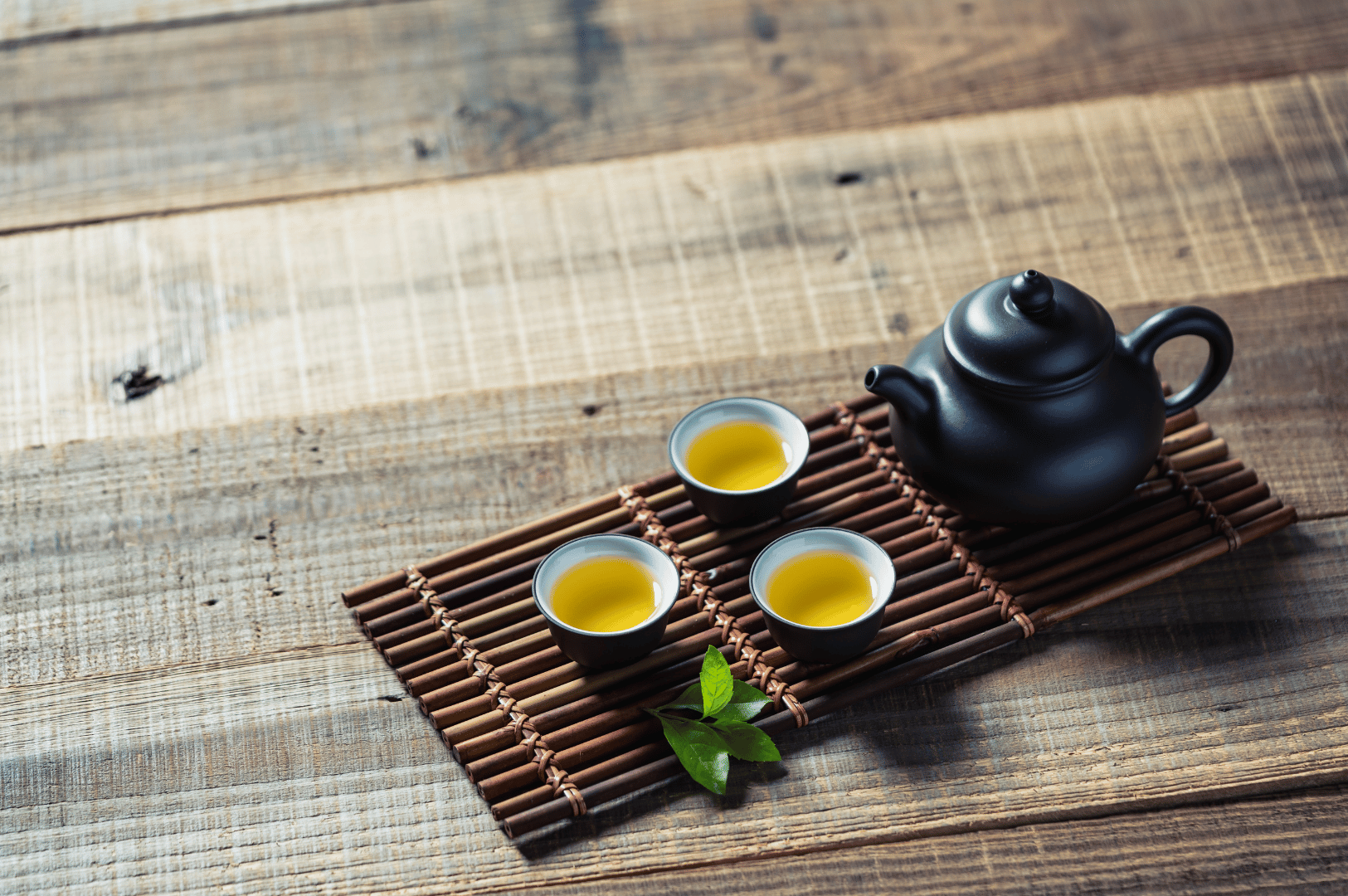 Chinese-Tea- (2) Black ceramic teapot and three teacups filled with yellow-green tea sit on a bamboo mat atop a wooden surface, accented by a sprig of fresh leaves—capturing the quiet refinement of Chinese tea rituals. This minimalist composition reflects tea’s 5,000-year journey from ancient healing practice to global cultural icon.