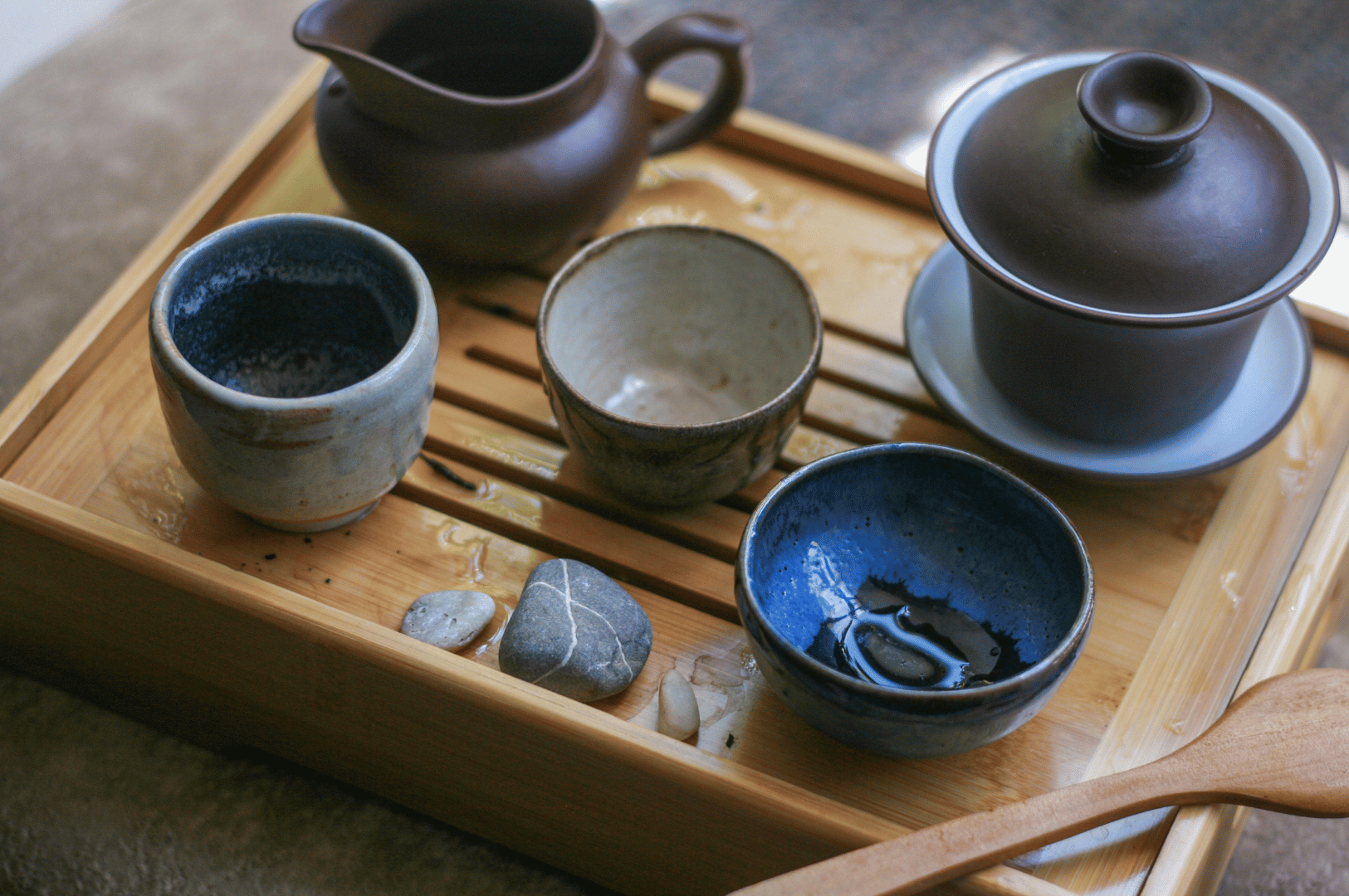 Chinese-Tea- (3) Traditional Chinese tea set arranged on a slatted wooden tray, featuring a brown teapot, lidded gaiwan, and three ceramic teacups in muted tones—blue, gray-white, and beige—beside decorative stones and a wooden spoon. The setup evokes the contemplative elegance of gongfu tea rituals, underscoring tea’s 5,000-year evolution from medicinal brew to global cultural emblem.