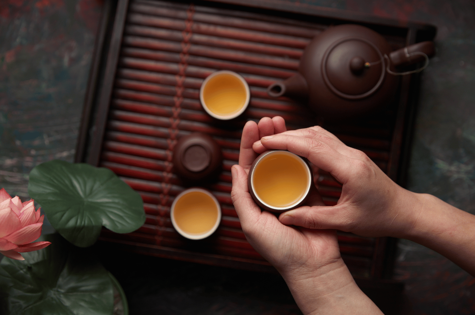 Chinese-Tea- (5) A person gently cradles a small cup of yellow tea above a wooden tray with slats, surrounded by a brown teapot, two additional filled cups, a green leaf, and a pink lotus flower—evoking the mindfulness and grace of Chinese tea ceremonies. This intimate moment reflects tea’s 5,000-year evolution from healing tradition to a global ritual of reflection and connection.