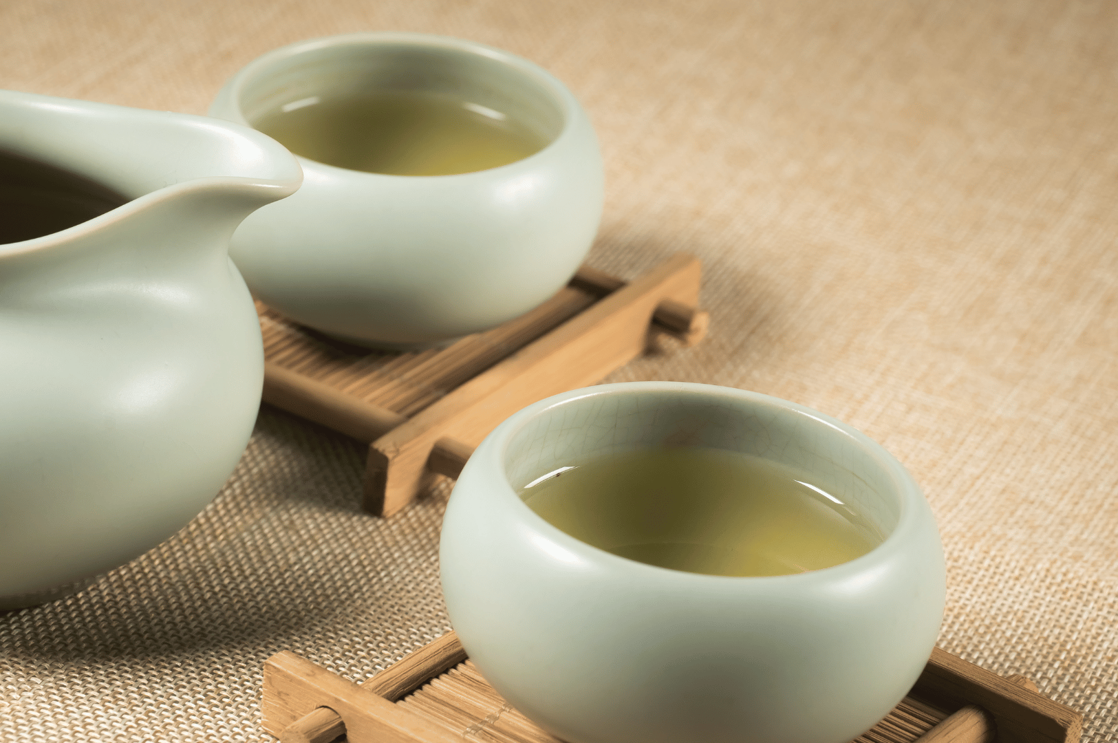 Chinese-Tea- (6) Two ceramic teacups filled with green tea rest on bamboo coasters beside a matching teapot, partially visible, against a warm beige fabric backdrop—evoking the understated beauty of Chinese tea traditions. This serene composition reflects tea’s 5,000-year journey from ancient healing ritual to a globally celebrated cultural practice.