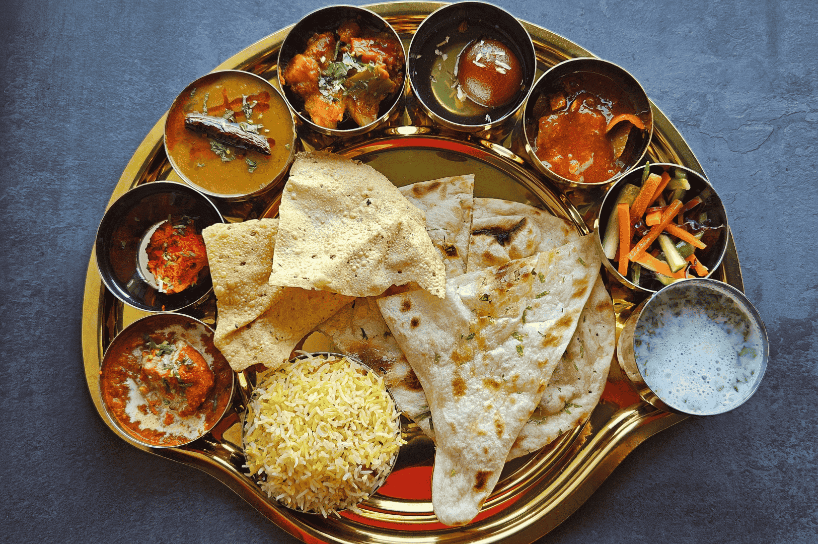 Gujarati-Thali- (2) Golden metal thali plate showcasing a vibrant Gujarati plant-based feast—yellow rice, spiced vegetable curries, creamy lentils, and tangy pickles arranged in ornate bowls. At the center, soft roti and crisp papad evoke warmth and tradition, while raita and herb garnishes add cooling contrast. A sensory celebration of Western India's culinary heritage, rich in color, texture, and flavor.
