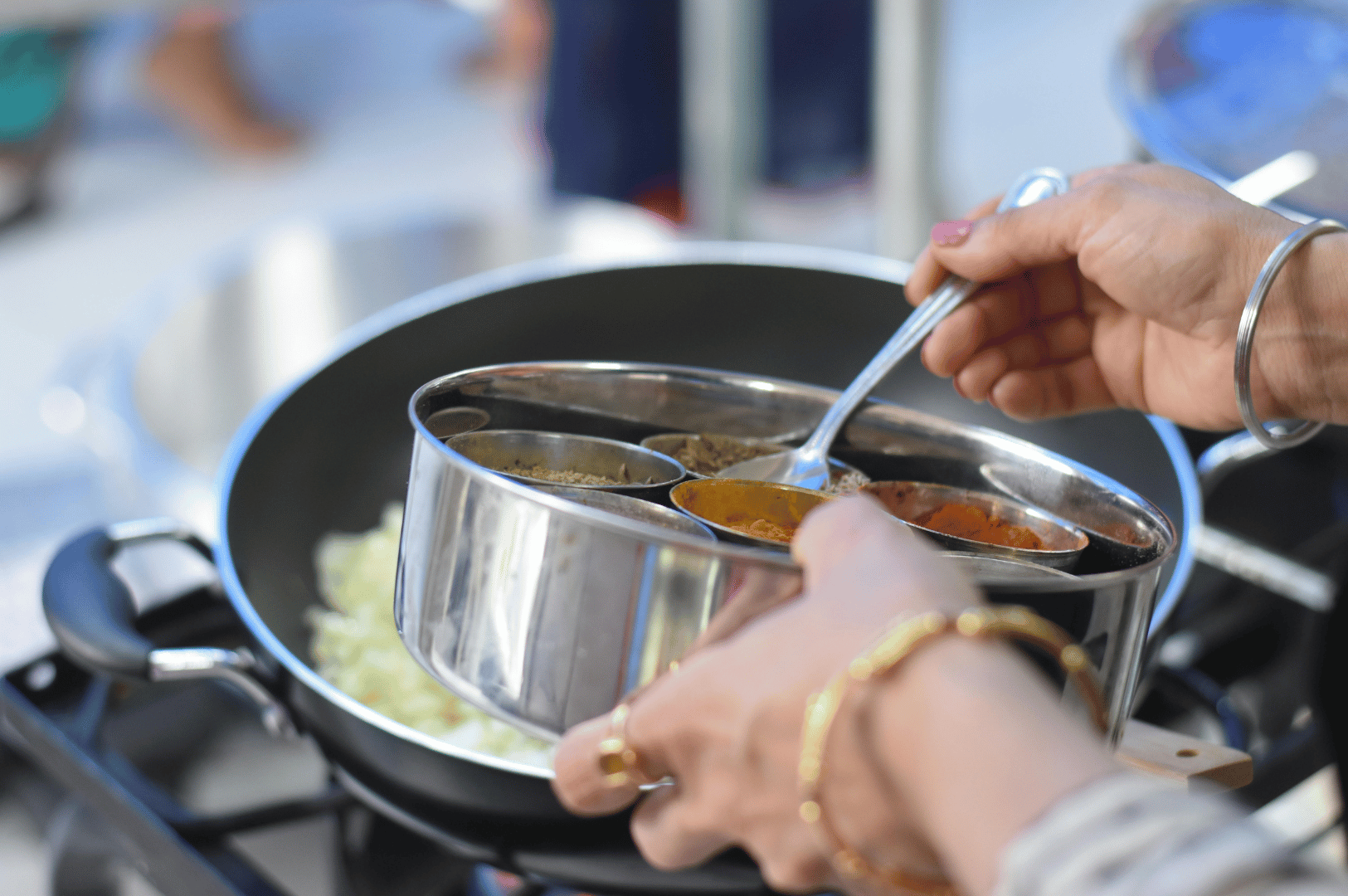 Gujarati-Thali- (4) Close-up of a home cook preparing Indian cuisine, scooping vibrant spices from a traditional stainless steel masala dabba into a pan of sautéed cabbage on a gas stove. Adorned with bangles and rings, the cook’s hands reflect cultural heritage and the intimate ritual of spice layering—an essential step in crafting the bold, plant-based flavors of a Gujarati thali.