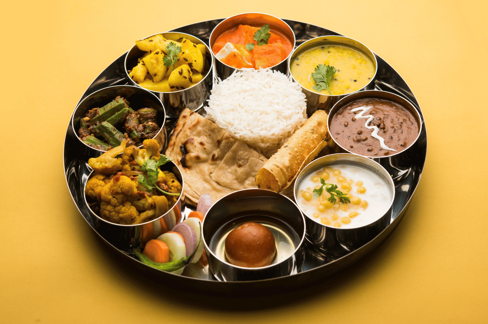 Gujarati-Thali- (5) Traditional Gujarati thali served on a round metal plate, centered with white rice and chapati, surrounded by vibrant plant-based dishes—potato curry with mustard seeds, lentil dal, okra stir-fry, aloo gobi, boondi raita, and paneer in tomato gravy. Garnished with cilantro and paired with papad, salad, and gulab jamun, this balanced feast celebrates Western India’s culinary diversity and vegetarian heritage.