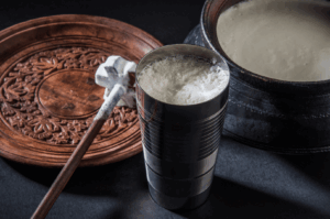 Traditional yogurt drink setup featuring a frothy metal cup of churned yogurt beside a wooden whisking tool and carved serving tray—evoking the artisanal preparation of Indian lassi and its regional variations across seasons.
