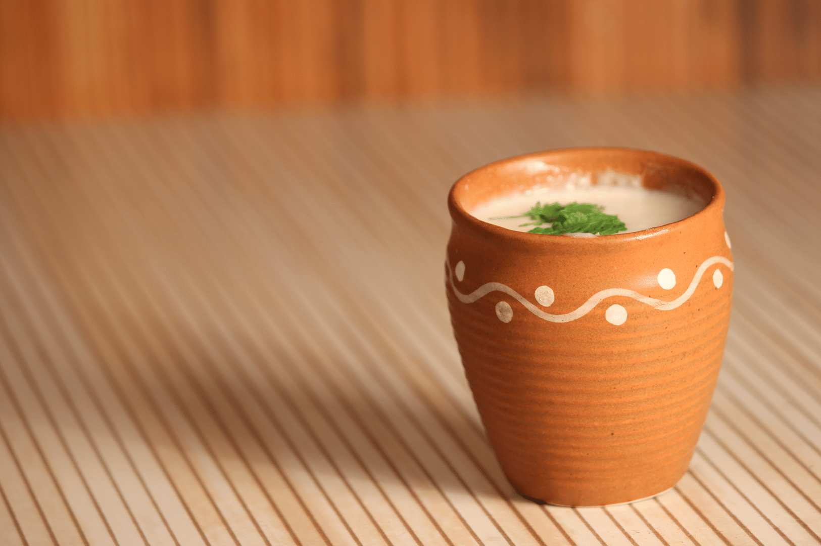 Indian-Lassi- (2) Rustic clay cup of creamy lassi garnished with fresh cilantro, set against a striped textile and wooden backdrop—highlighting India’s seasonal yogurt traditions and regional serving aesthetics