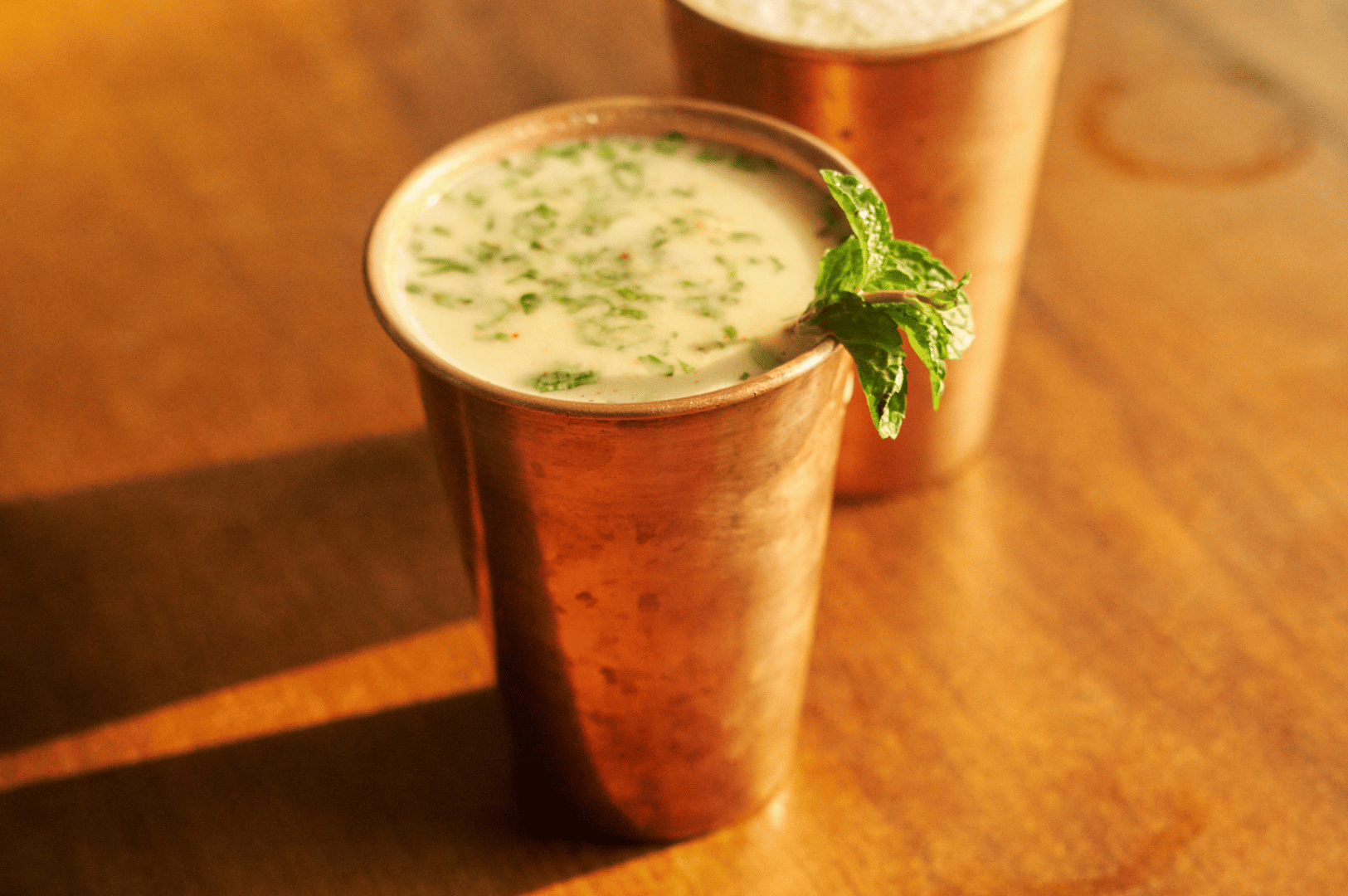 Indian-Lassi- (3) Pair of copper tumblers filled with herb-garnished masala chaas, set on a wooden surface—celebrating India’s spiced yogurt traditions and the tactile elegance of regional drinkware.