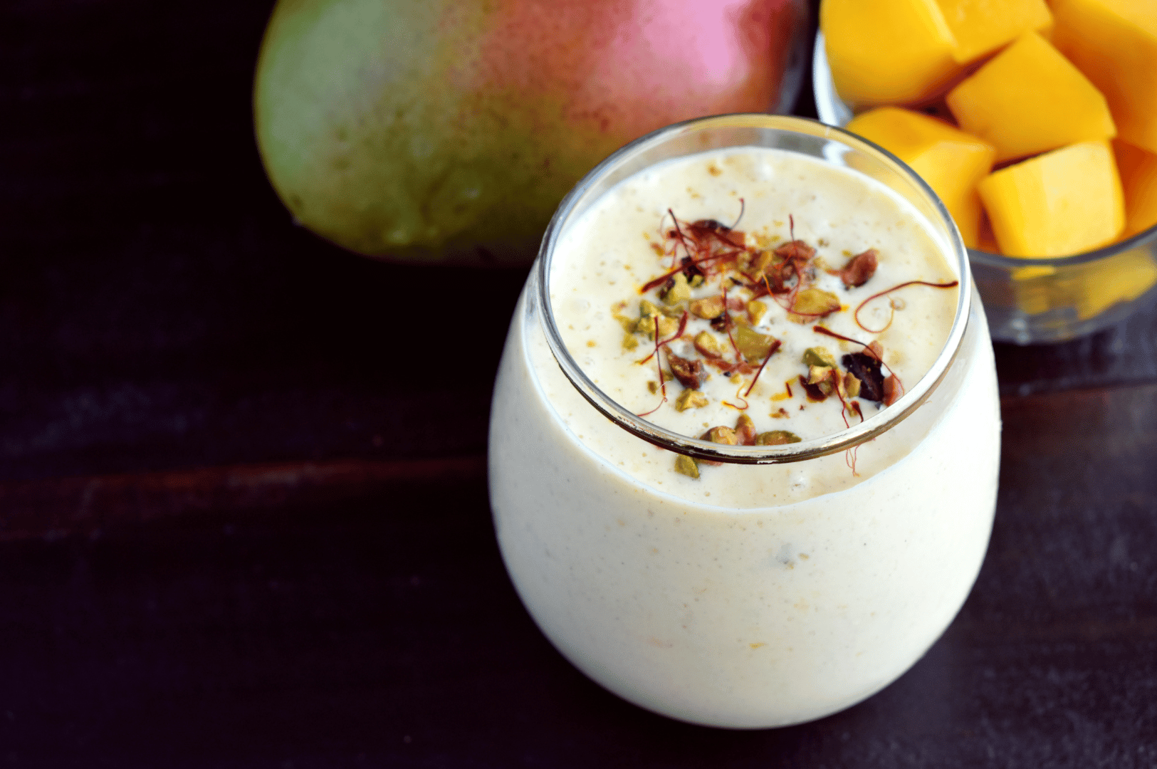 Indian-Lassi- (6) Glass of mango lassi topped with pistachios and saffron, framed by fresh mango and diced fruit—celebrating India’s sun-kissed yogurt traditions and the golden richness of seasonal ingredients.