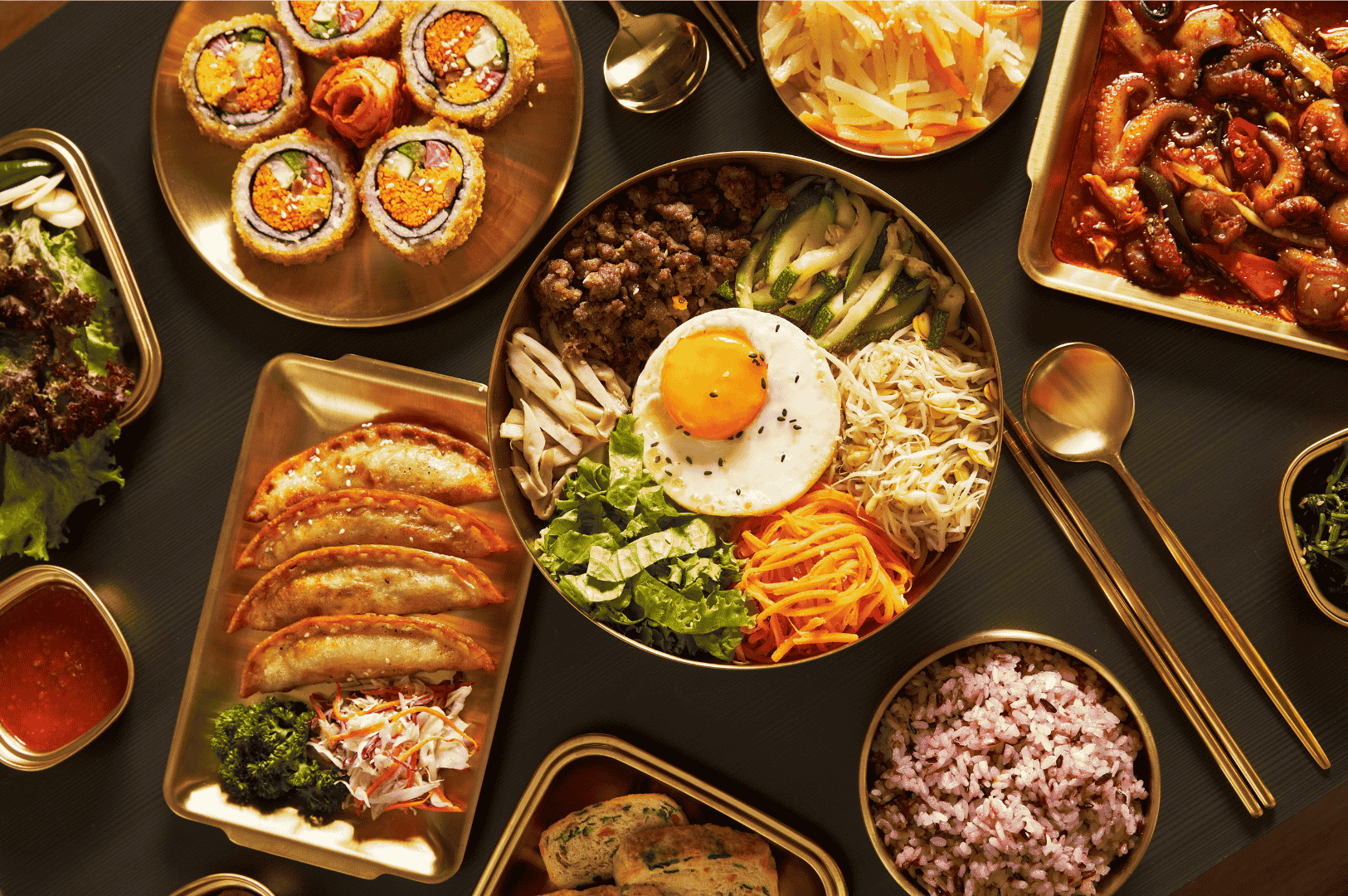 Jeonju- (1) A vibrant spread of Korean side dishes and bibimbap ingredients arranged in gleaming gold trays on a patterned table, highlighting Jeonju’s rich culinary presentation.