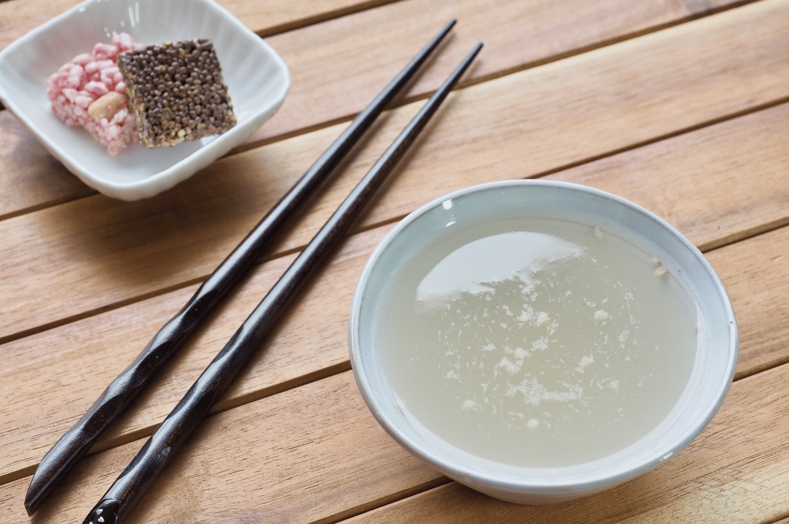 Sikhye- (2) A ceramic bowl of translucent Sikhye sweet rice punch sits on a warm wooden surface, its clarity revealing floating grains of cooked rice. Beside it, twisted dark chopsticks add a sculptural touch, while a small white dish in the background offers puffed rice and sesame snacks—seasonal accompaniments that evoke spring renewal and communal sharing. The composition blends rustic elegance with traditional Korean textures, deepening the sensory narrative of Sikhye through the seasons.