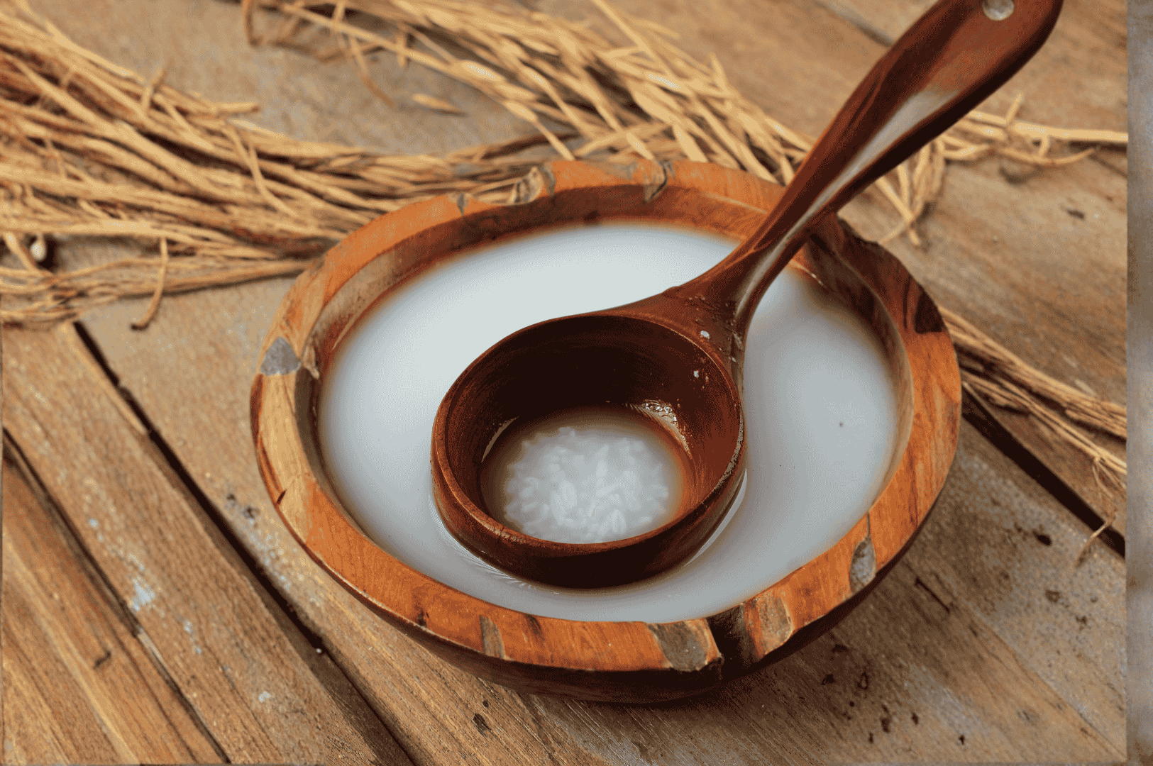 Sikhye- (3) A wooden bowl of creamy Sikhye sweet rice punch sits atop a matching wood surface, its ladle cradling a few tender rice grains in a pool of milky liquid. Behind it, dried rice stalks fan out like a harvest offering, grounding the scene in autumnal warmth and ancestral memory. The earthy textures and soft lighting evoke the cyclical grace of Korean tradition, where Sikhye bridges nourishment and ritual through the changing seasons.