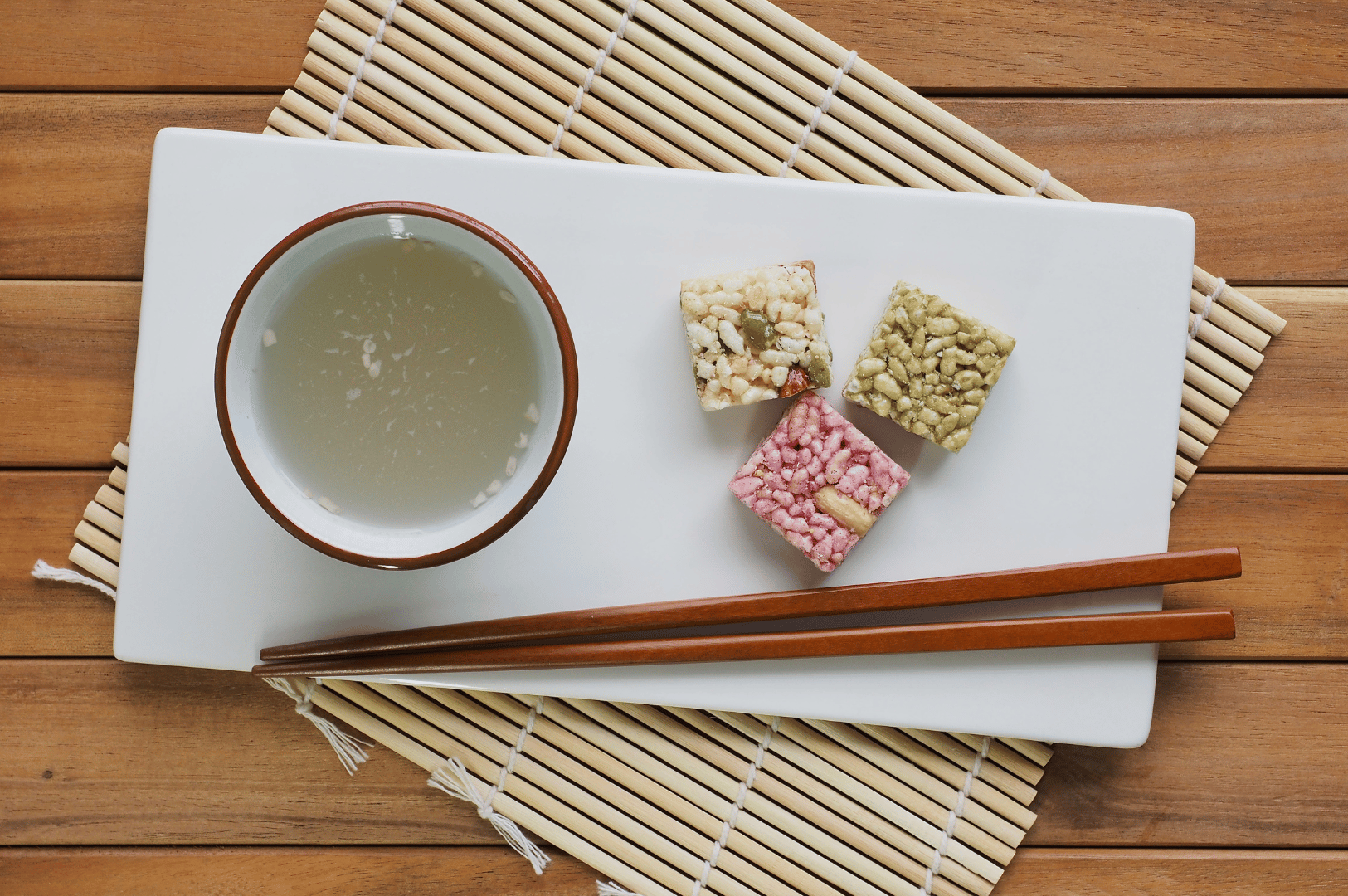 Sikhye- (4) A white rectangular plate on a bamboo mat showcases a bowl of translucent Sikhye sweet rice punch, paired with three puffed rice snacks in soft pastel hues—white, pink, and green. Wooden chopsticks rest neatly beside the bowl, echoing the clean lines and seasonal elegance of Korean springtime rituals. The composition celebrates renewal and balance, inviting readers into the gentle joy of sharing Sikhye across generations.