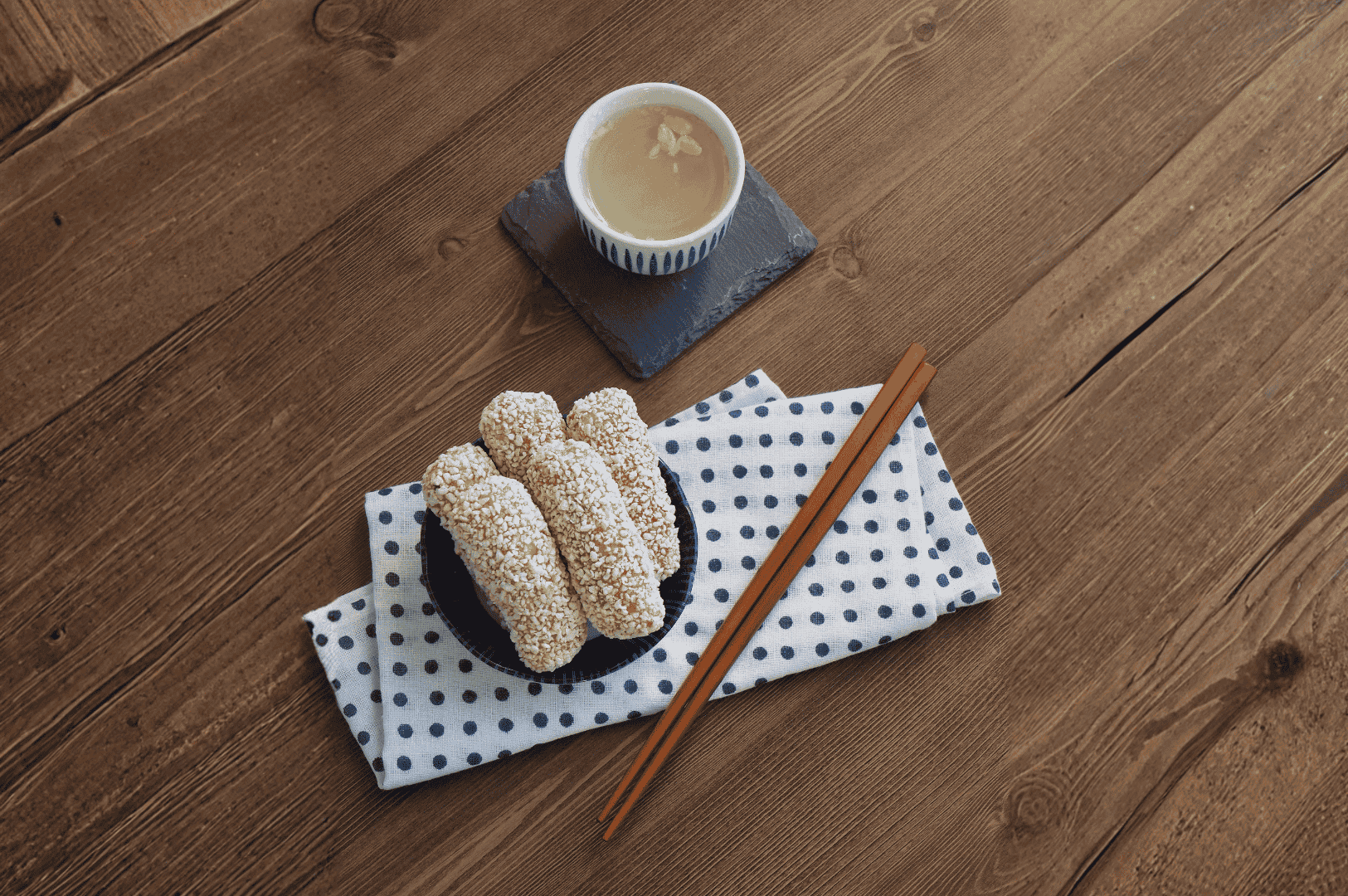Sikhye- (5) A small bowl of sesame-coated rice snacks rests on a wooden surface, accompanied by diagonally placed wooden chopsticks atop a white cloth with black polka dots. Above, a warm cup of light-colored tea sits on a dark coaster, adding a quiet elegance to the scene. The minimalist arrangement evokes winter introspection and Korean hospitality, where Sikhye and its seasonal pairings offer comfort and connection.