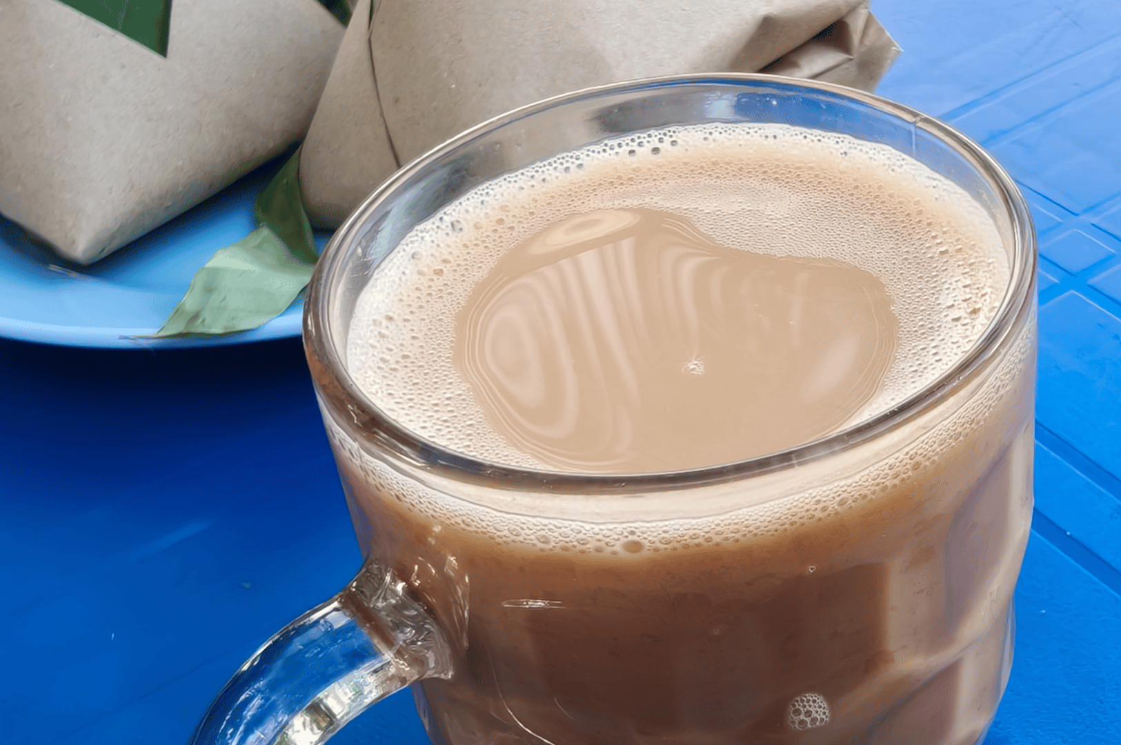 A cup of pulled milk tea in a glass mug on a blue table, capturing the creamy froth typical of Malaysian teh tarik.