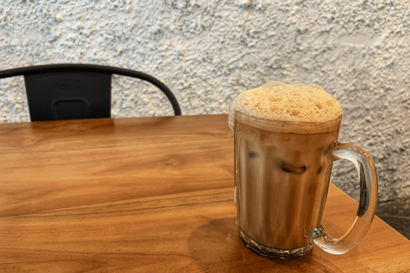 A tall glass of teh tarik on a wooden table with a paper straw, condensation beading on the glass, evoking Malaysia’s beloved teh tarik in a chilled form.