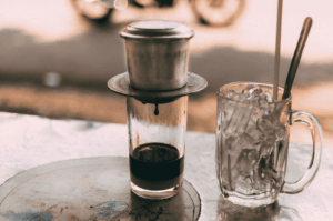 Traditional Vietnamese coffee brewing in progress: a metal phin filter slowly drips rich, dark coffee into a glass, while a separate iced mug awaits for mixing. Set against a blurred street scene, the image captures the ritualistic calm and urban vibrancy of Ca Phe Sua Da culture—where patience, contrast, and sensory anticipation define the experience.